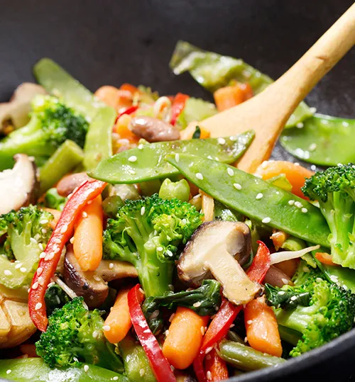 Gemüse-Stir-Fry in schwarzem Wok mit Holzkelle: Brokkoli, Zuckerschoten, rote Paprika und Pilze