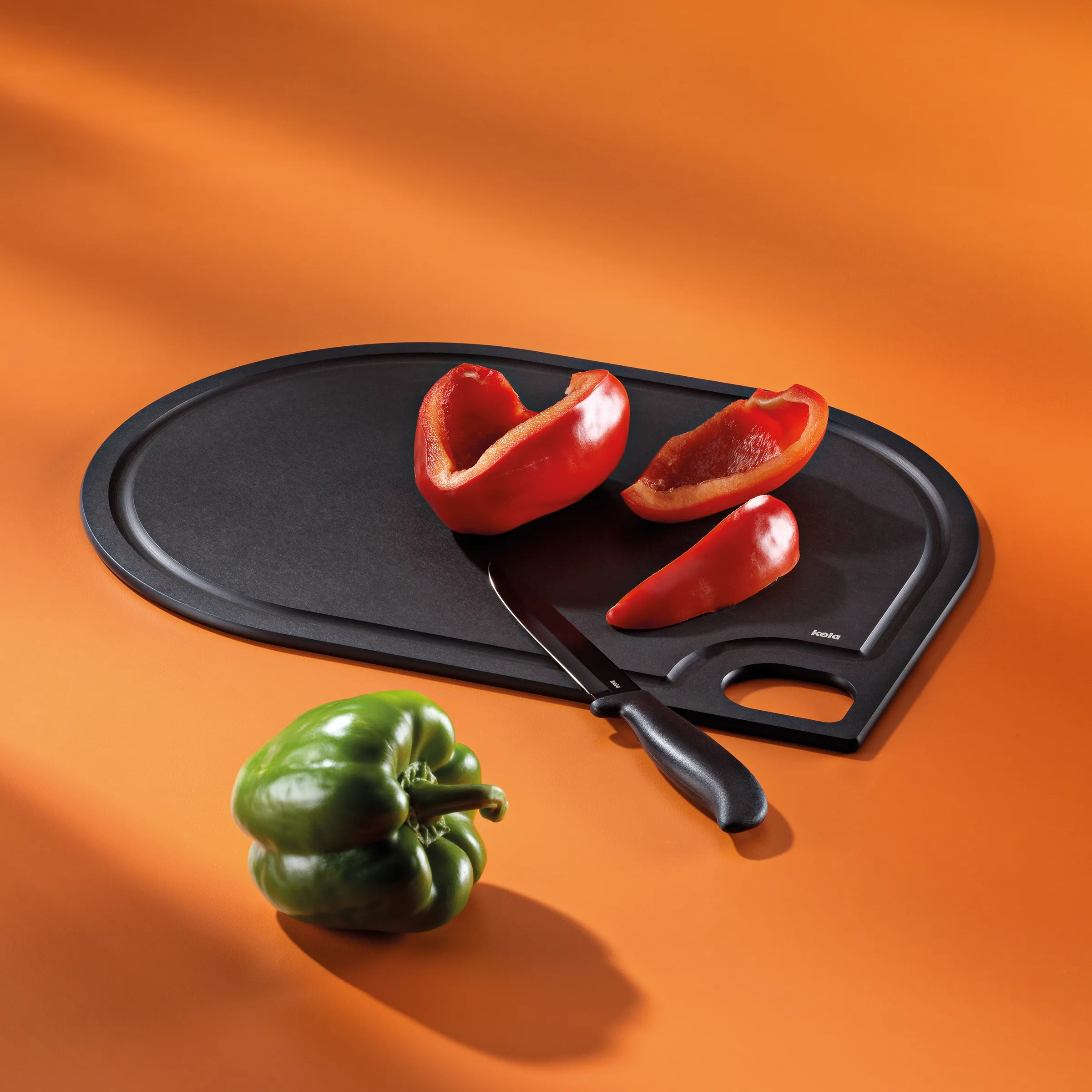 Schneidebrett-Set mit Messer und Paprika - Küchenzubehör Schwarzes Schneidebrett-Set mit Messer, rote und grüne Paprika auf orangefarbenem Hintergrund