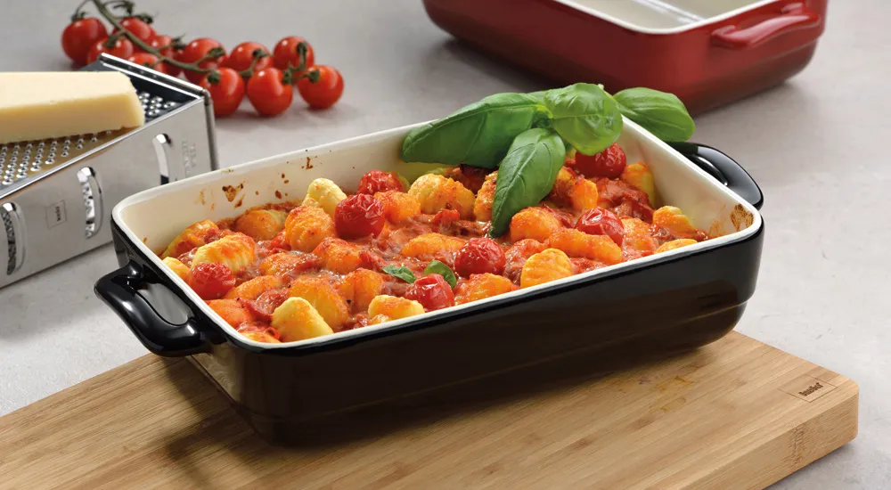 Gnocchi in Tomatensoße - Italienische Auflaufform Schwarze Ofenform mit Gnocchi in Tomatensoße, garniert mit frischem Basilikum auf Holzbrett, umgeben von Tomaten und Käsereibe