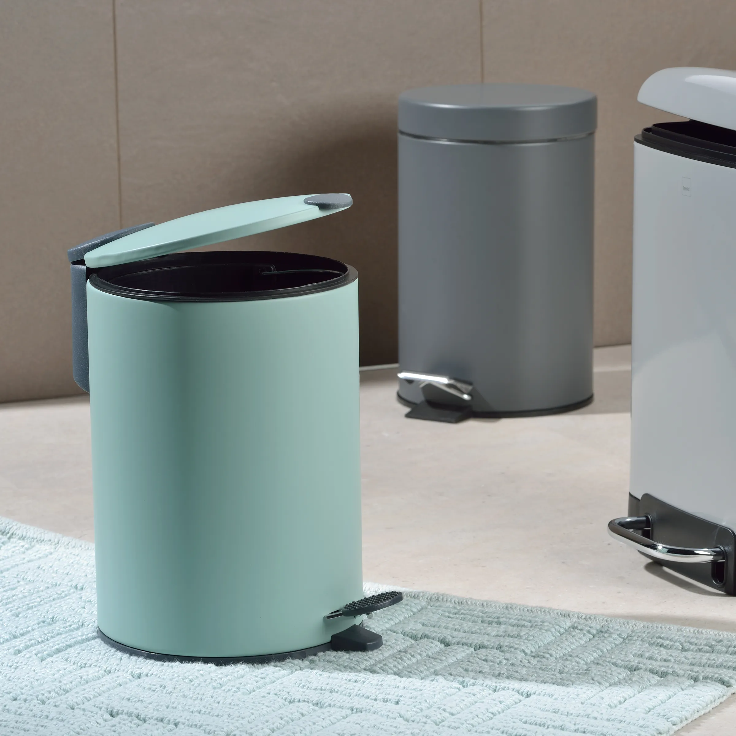 Moderne Tretmülleimer in verschiedenen Farben - Badezimmer Abfalleimer mit Fußpedal Drei moderne Tretmülleimer in Mint, Grau und Weiß auf Badezimmerteppich vor beiger Wand