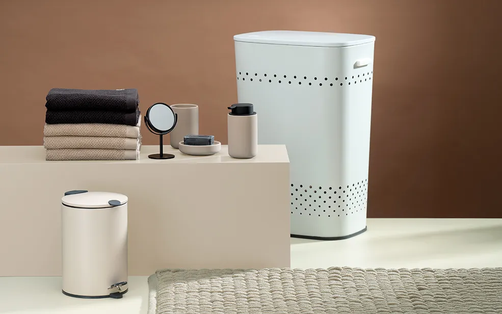 Badezimmer-Accessoires Set mit Wäschekorb und Mülleimer Badezimmer-Set mit weißem Wäschekorb, Mülleimer, Behältern und gestapelten schwarzen sowie beigen Handtüchern auf neutralem Hintergrund.