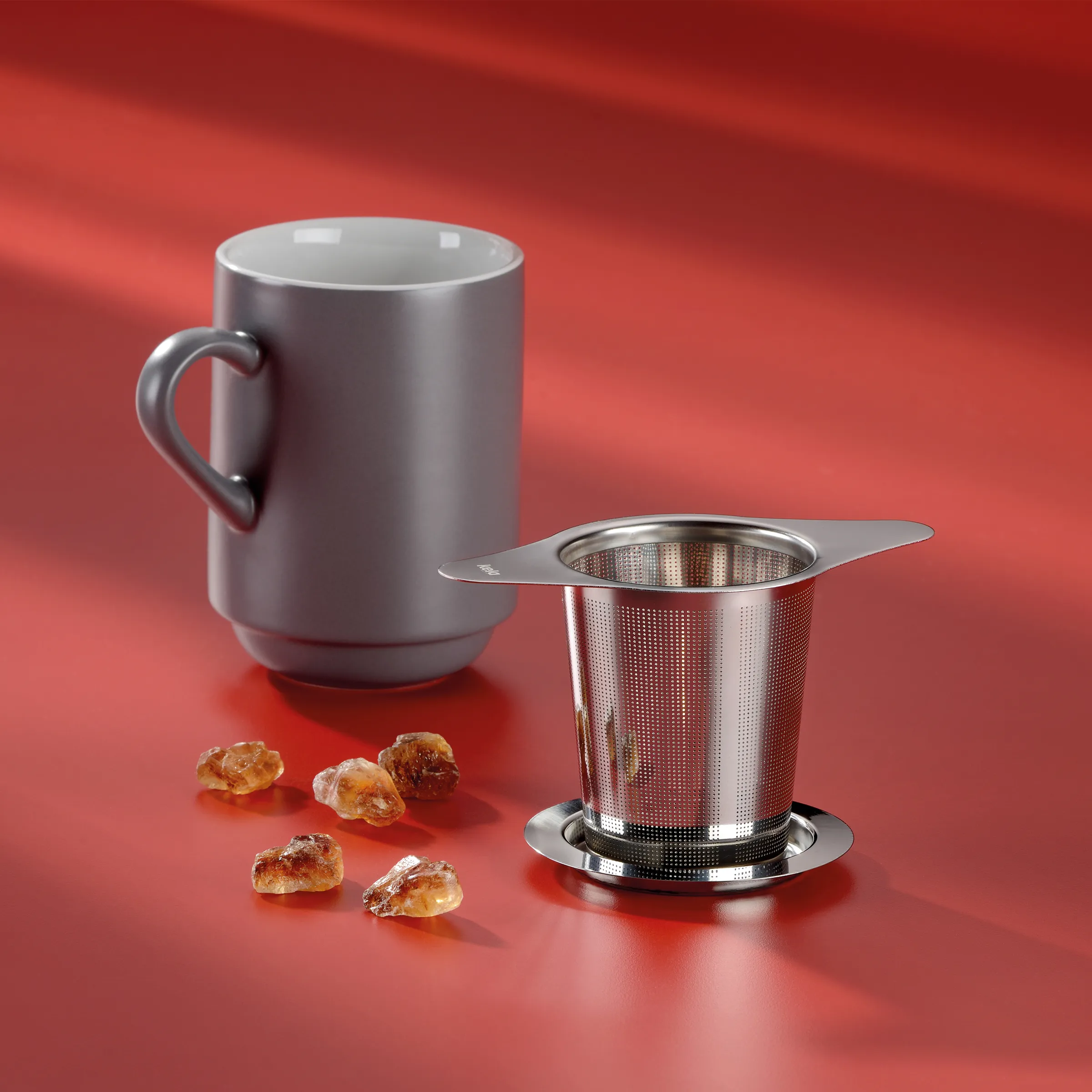 Graue Keramiktasse mit Henkel und Edelstahl-Teesieb auf rotem Hintergrund, daneben Bernsteinstücke