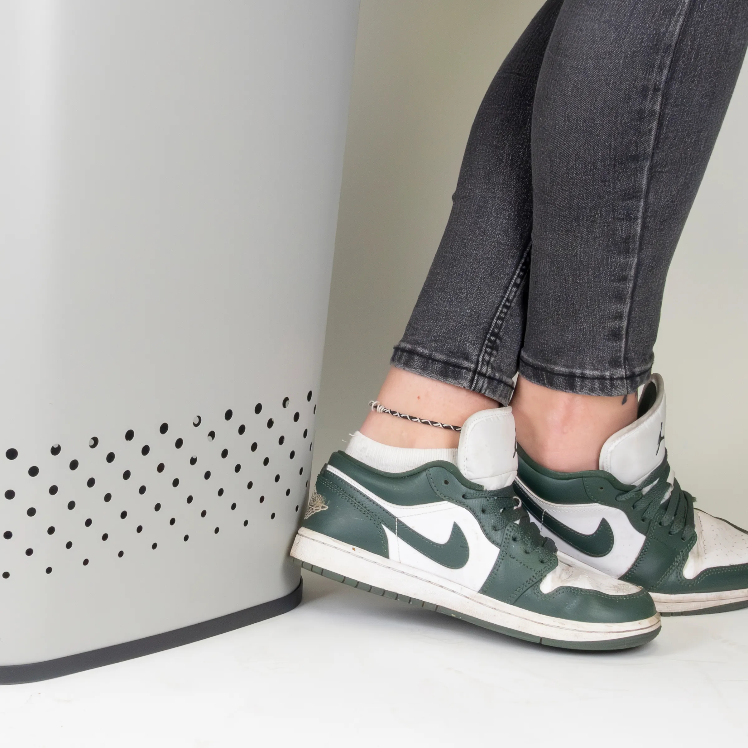 Person trägt grüne und weiße Jordan 1 High Sneaker mit grauer Jeans und weißem Socken vor perforierter weißer Wand