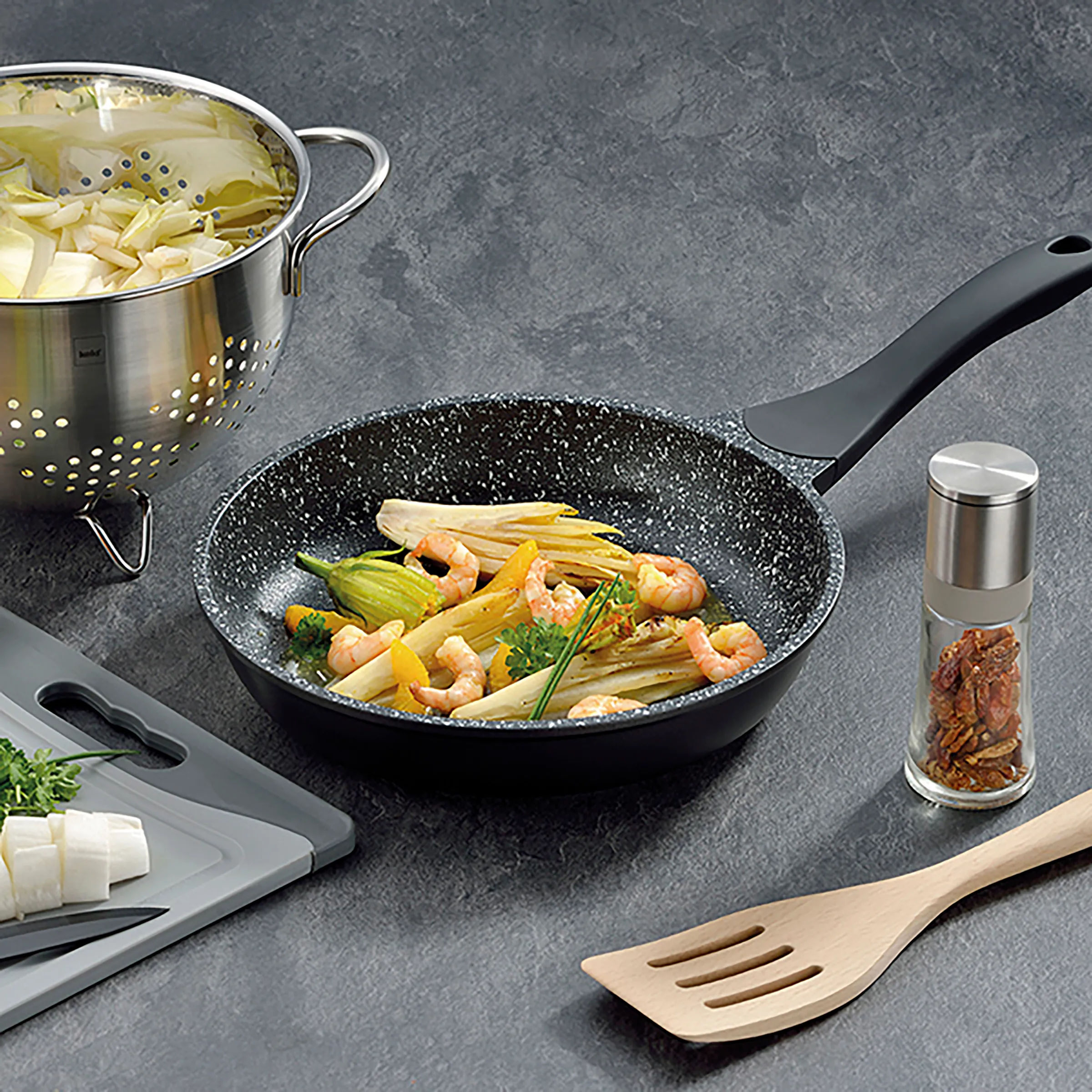 Wok-Pfanne antihaftbeschichtet mit Garnelen-Gemüse-Rezept Schwarze Antihaft-Wok-Pfanne mit gebratenen Garnelen und Gemüse, daneben Nudelsieb, Kochlöffel und Gewürzstreuer auf grauem Untergrund.