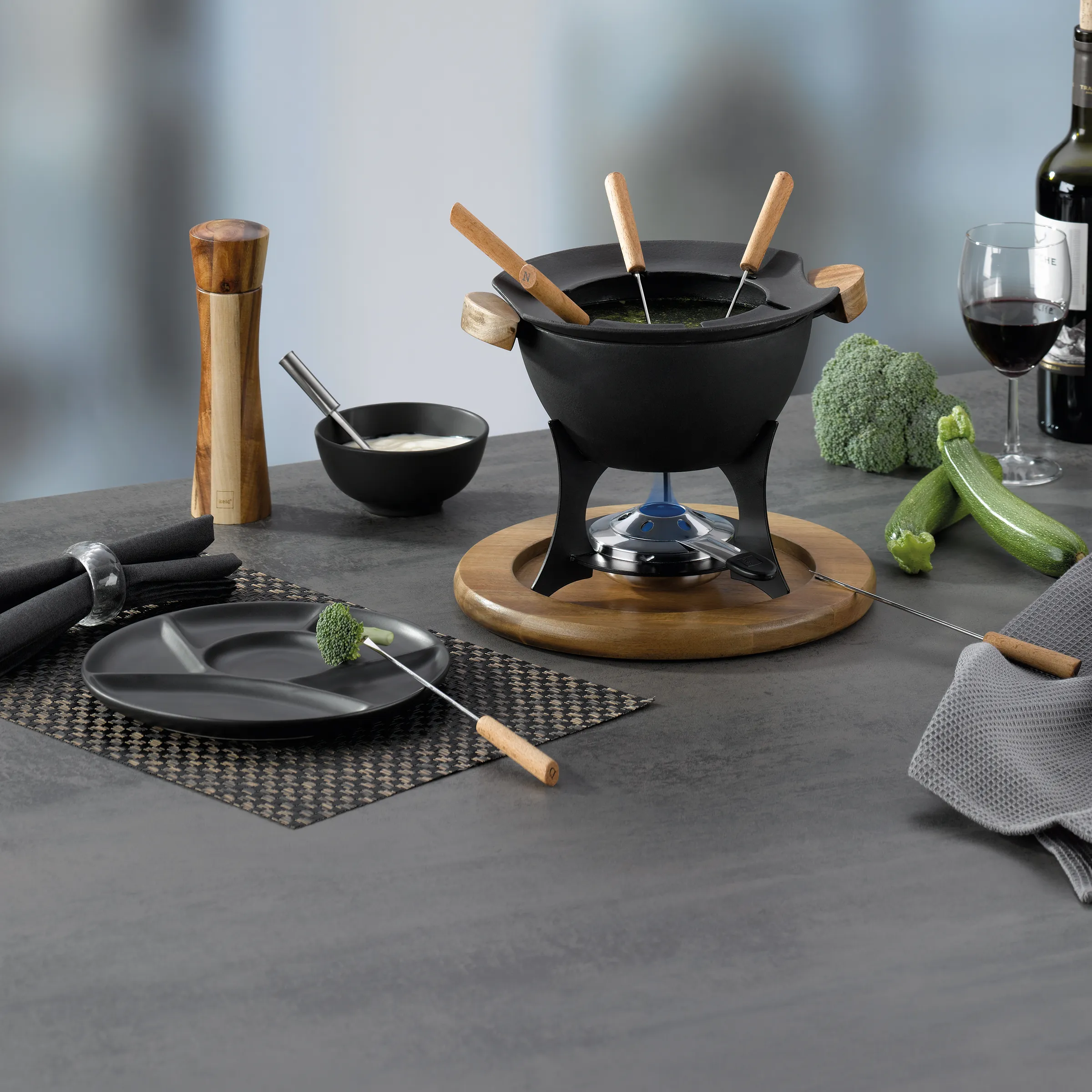 Fondue-Set schwarz mit Holzgriffen - Käsefondue Geschirr Set Schwarzes Fondue-Set mit Holzgriffen auf rundem Brett, umgeben von Käseschüssel, Gemüse und Rotwein