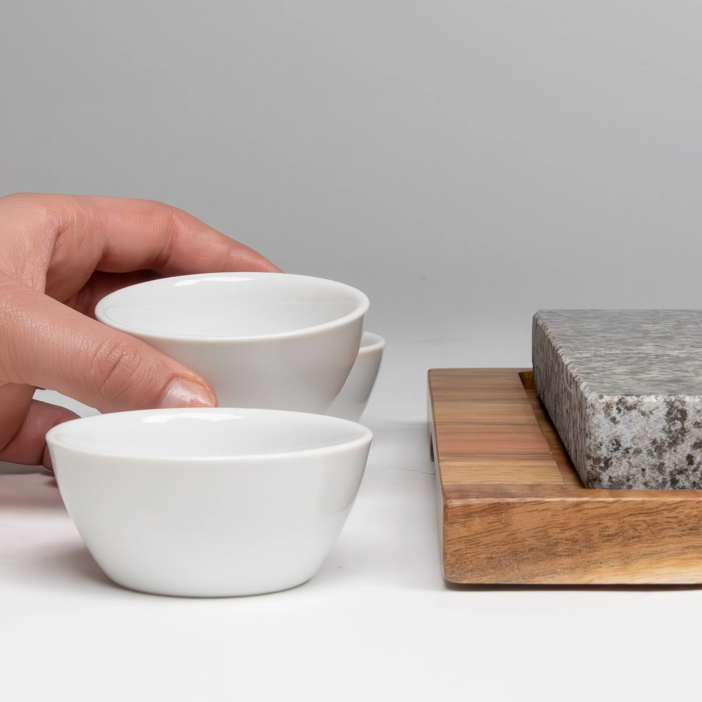 Weiße Porzellanschalen und Mörserset - Küchenausstattung Hand hält kleine weiße Porzellanschale neben Holzbrett mit Steinblock und Granitmörser auf grauem Hintergrund
