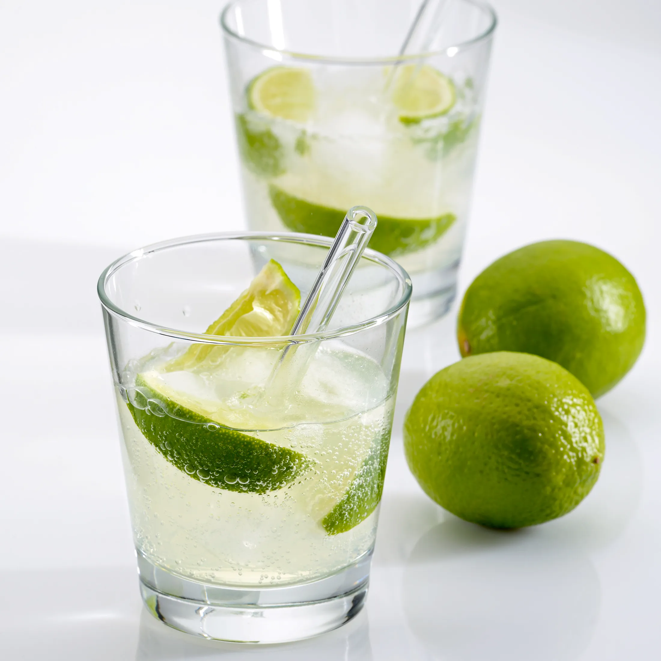 Erfrischendes Limetten-Getränk mit Eis und Glasstrohhalm - Sommergetränk Zwei Gläser mit sprudelndem Getränk, Limettenscheiben und Eiswürfeln, daneben zwei frische Limetten