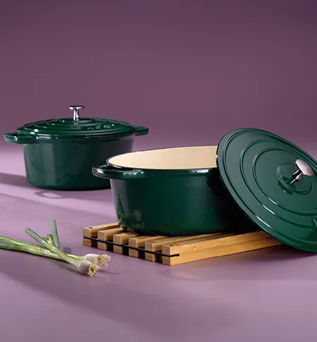 Zwei grüne Gusseisen-Bräter mit Deckel auf violettem Hintergrund, daneben Holzuntersetzer und Lauch