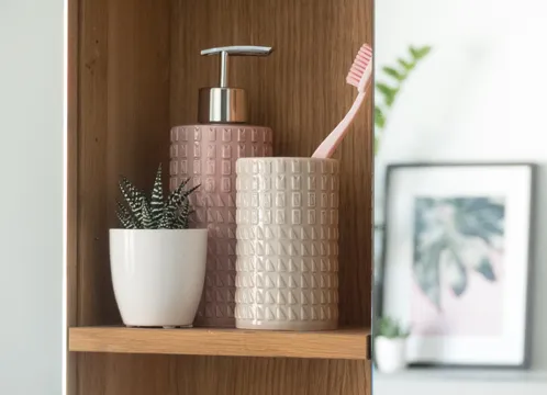 Badezimmer-Regal mit rosa Seifenspender, weißem Blumentopf, Zahnbürste und Pflanze auf Holzbrett