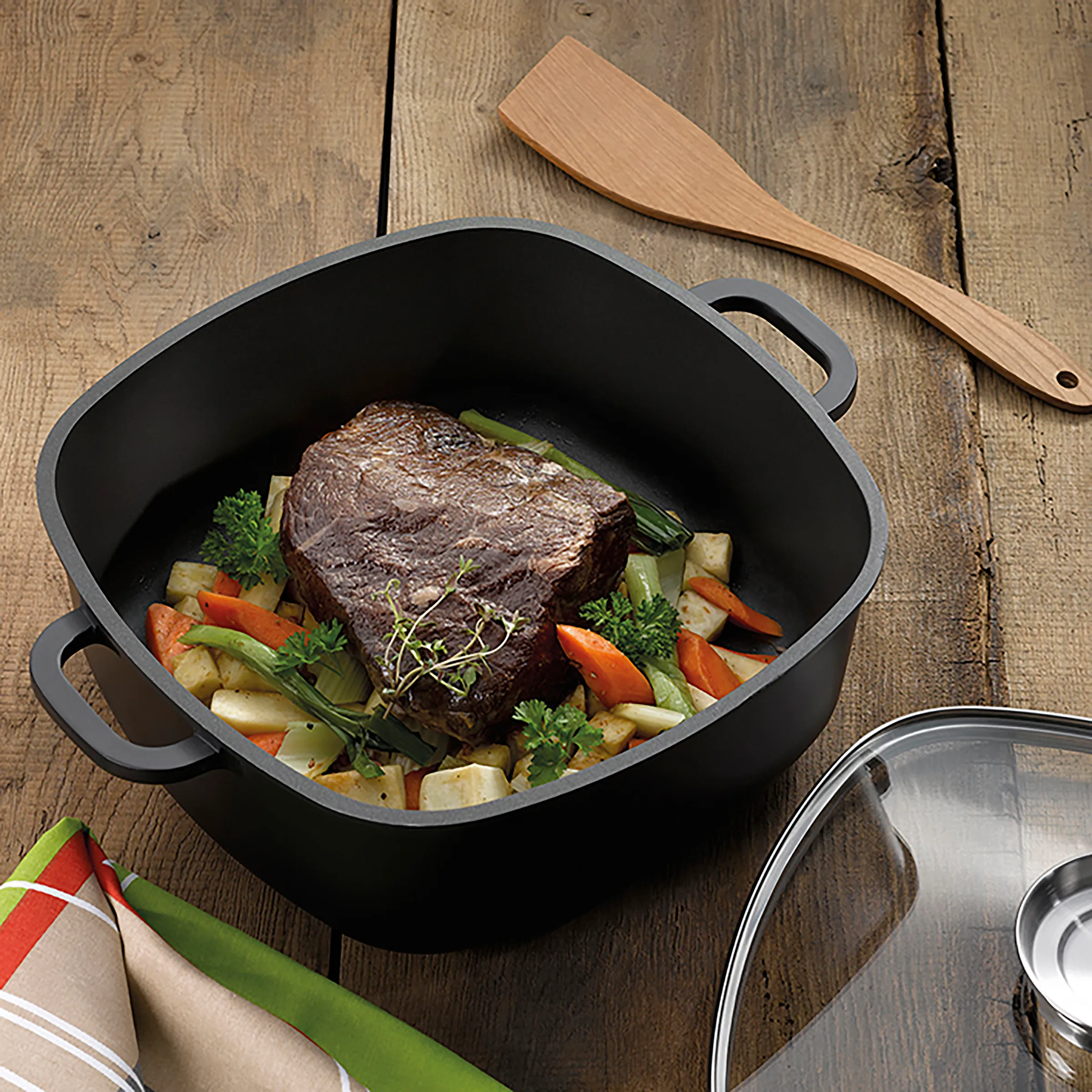 Schmortopf Gusseisen: Rinderbraten mit Gemüse zubereiten Schwarzer Schmortopf mit Deckel und Griffen auf Holztisch mit Rinderbraten, Gemüse und Kräutern