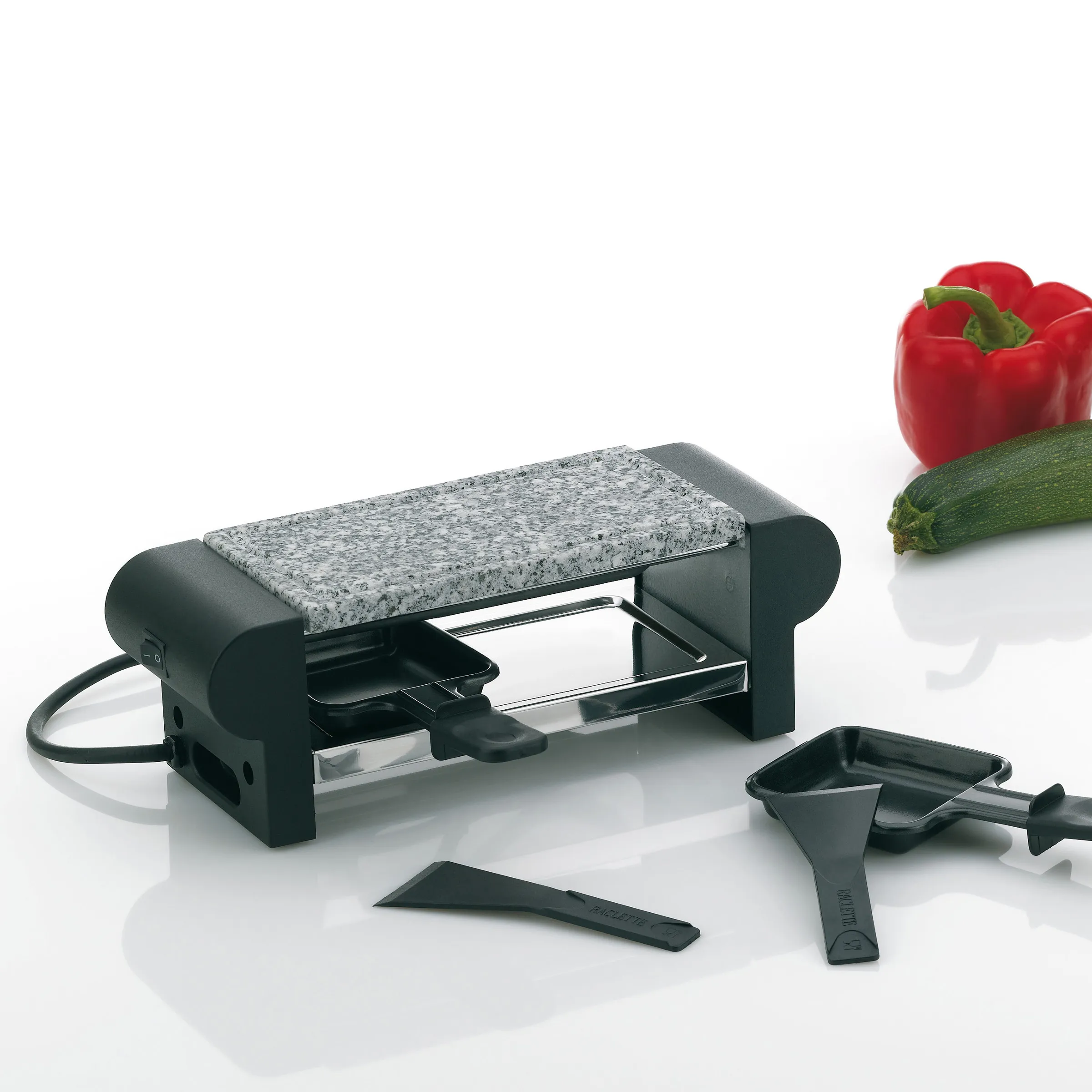 Raclette-Grill für 2 Personen mit Naturstein und Antihaft-Pfännchen Raclette-Grill für 2 Personen mit Granitstein, zwei Pfännchen und Schaber auf weißem Hintergrund mit Paprika
