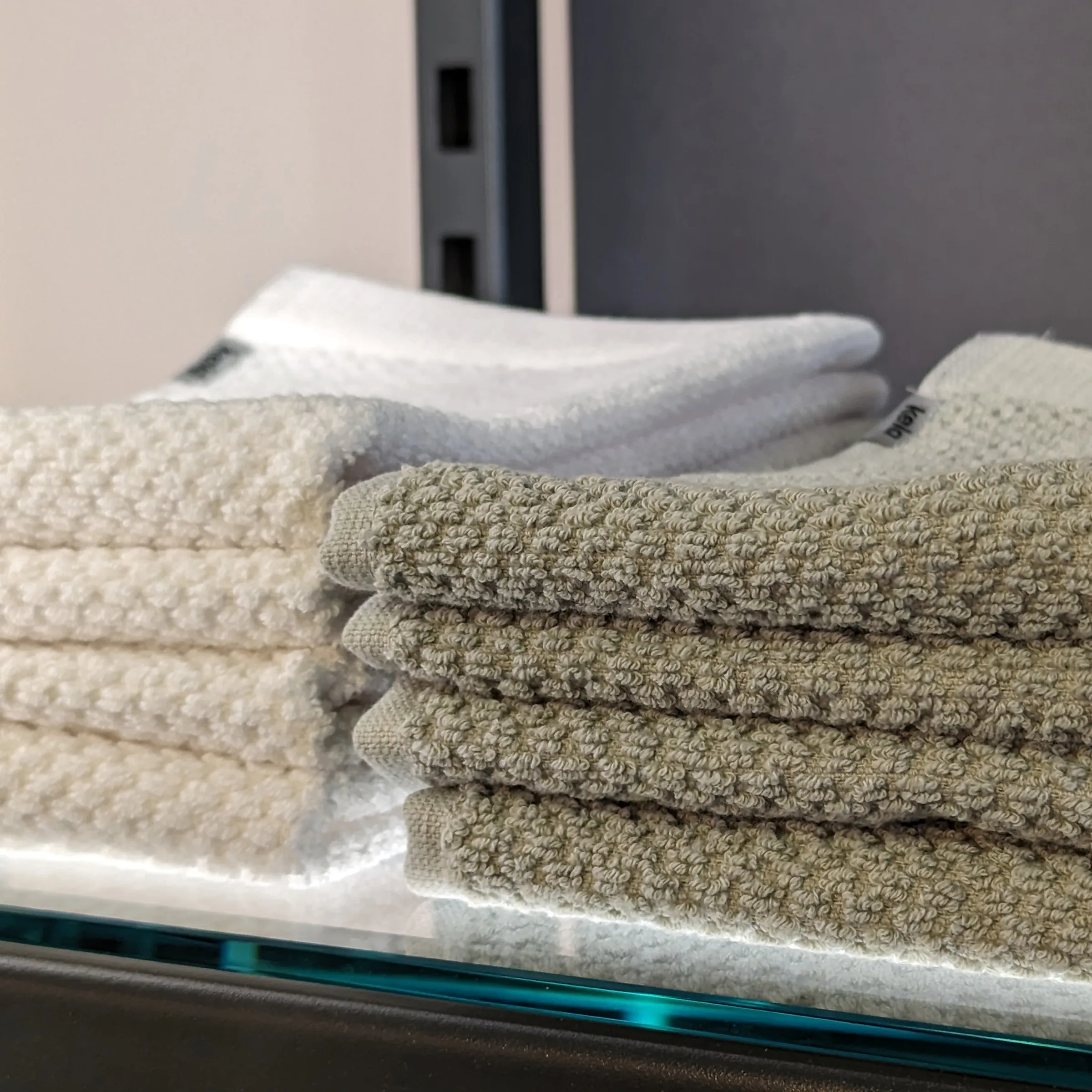 Premium Handtücher Set Beige Weiß für Badezimmer Gestapelte Handtücher in Beige und Weiß auf Glas, dahinter moderne grau-schwarze Badezimmerausstattung
