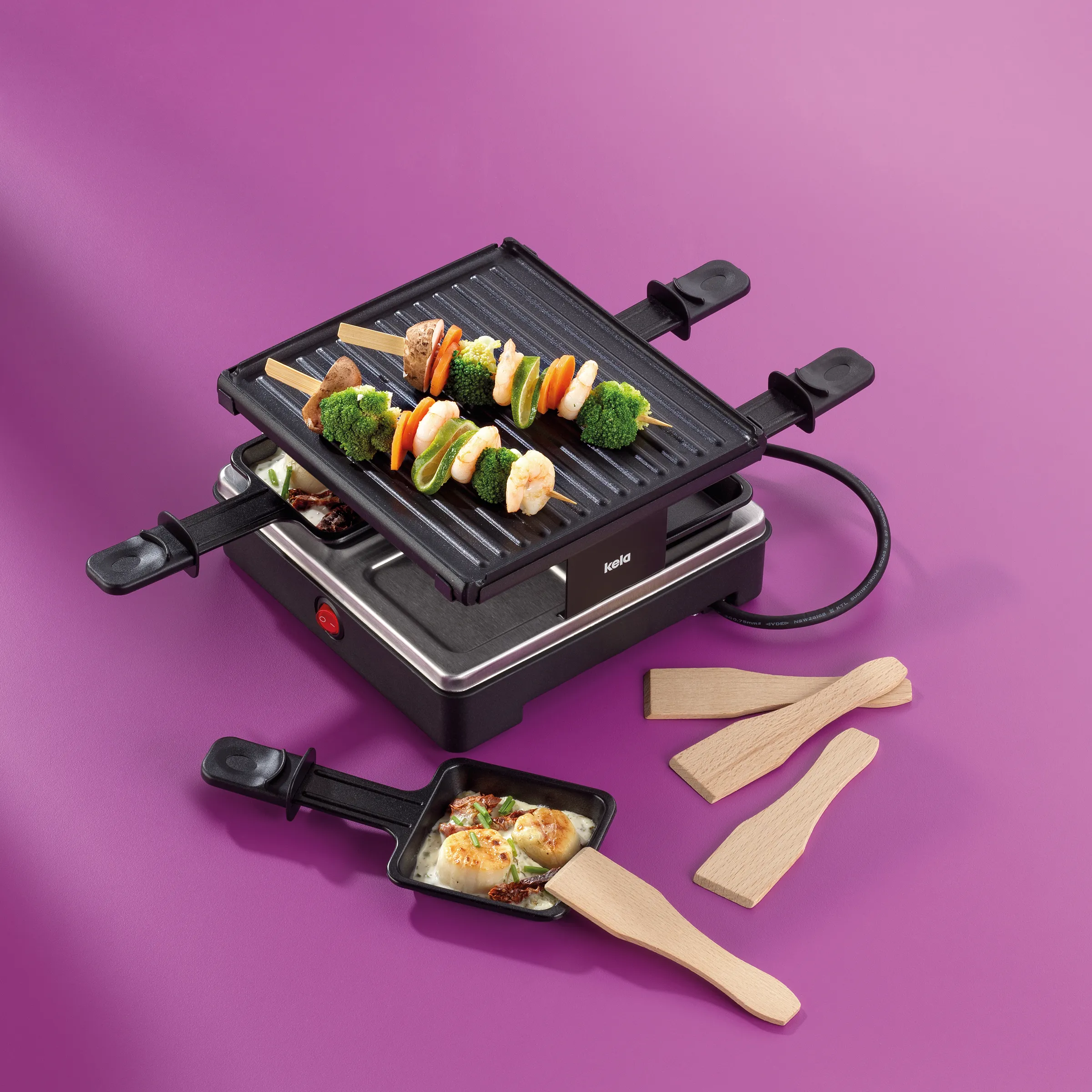 Kela Raclette-Grill mit Gemüsespießen auf gegrillter Platte und kleinen Pfännchen, Holzspatel auf violettem Hintergrund
