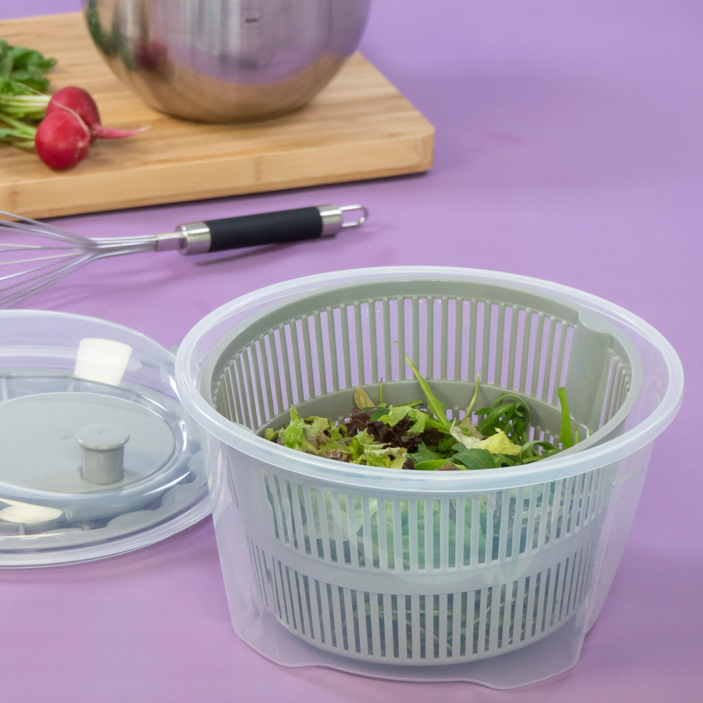 Salatschleuder mit frischem Salat - Küchenhelfer zum Trocknen von Blattsalat Salatschleuder mit frischem grünem Blattsalat auf lila Untergrund mit Schneebesen und Schneidebrett