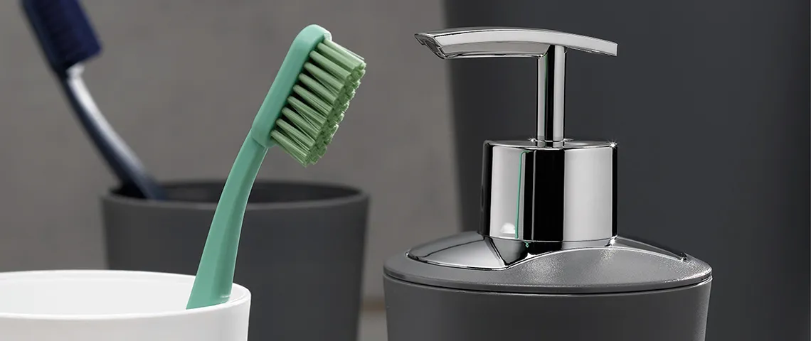 Badezimmer Accessoires Set mit Zahnbürste und Seifenspender Badezimmer-Accessoires: grüne Zahnbürste in weißem Becher, silberne Seifenspender-Pumpflasche und weitere Hygieneartikel auf grauem Hintergrund.