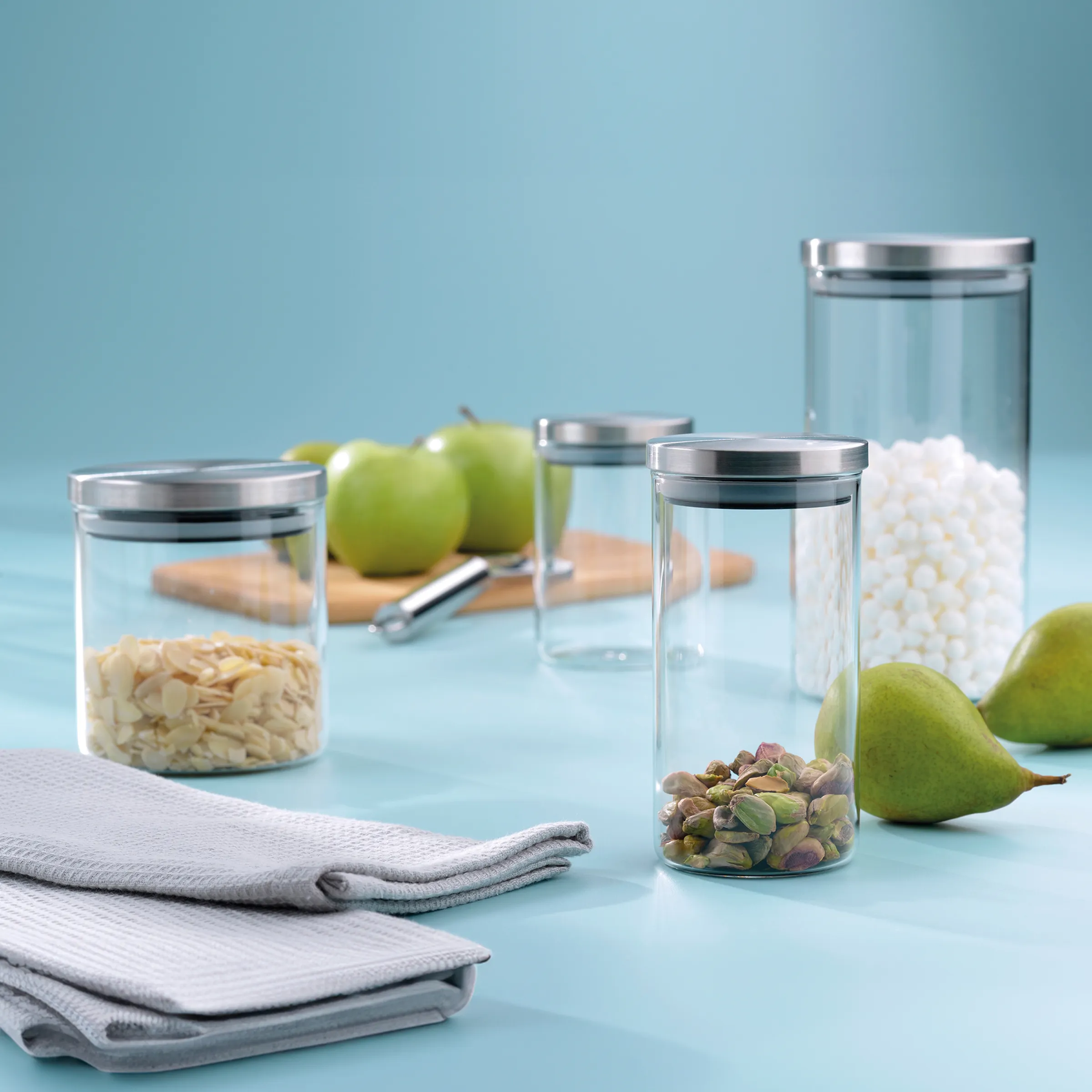Vorratsgläser mit Lebensmitteln - Aufbewahrungsbehälter für Küche Vier Glasdosen mit Metalldeckeln gefüllt mit Haferflocken, Pistazien und anderen Zutaten neben grünen Äpfeln und Birnen auf hellblauem Hintergrund