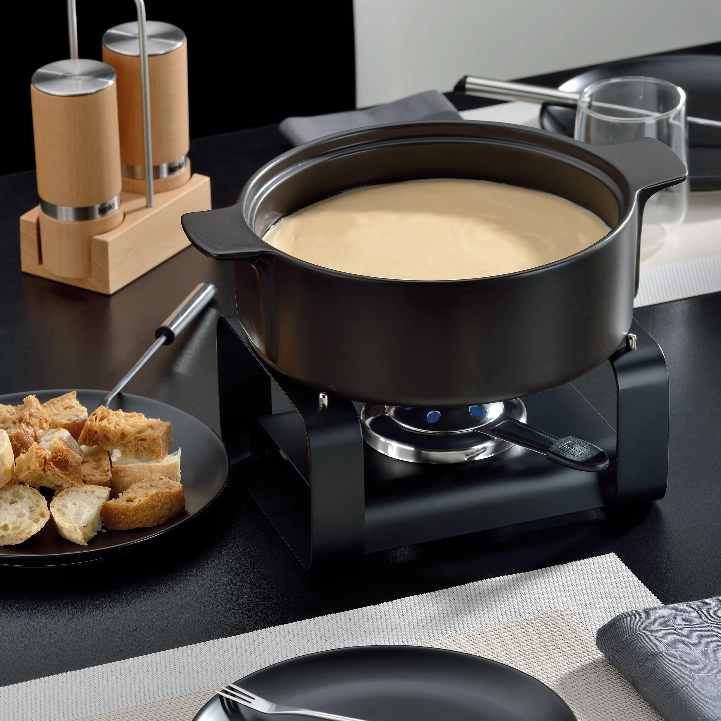 Käsefondue-Set schwarz mit Rechaud und Zubehör - moderne Fondue-Garnitur Schwarzes Käsefondue-Set mit cremigem Käse, Brotwürfeln auf einem Teller und Gewürzstreuer auf dunkler Oberfläche