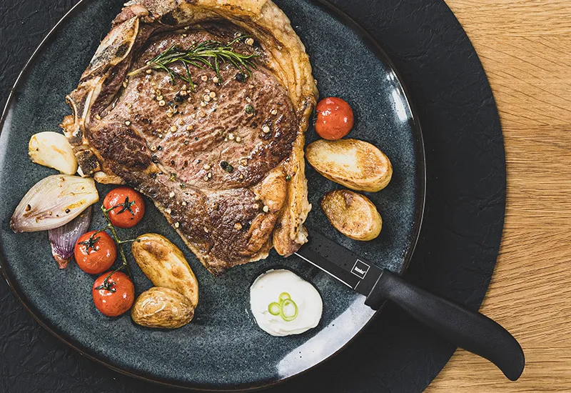 Gebratenes Steak mit Rosmaringarnitur, röstigen Kartoffeln, Tomaten und Zwiebeln in einer schwarzen Pfanne