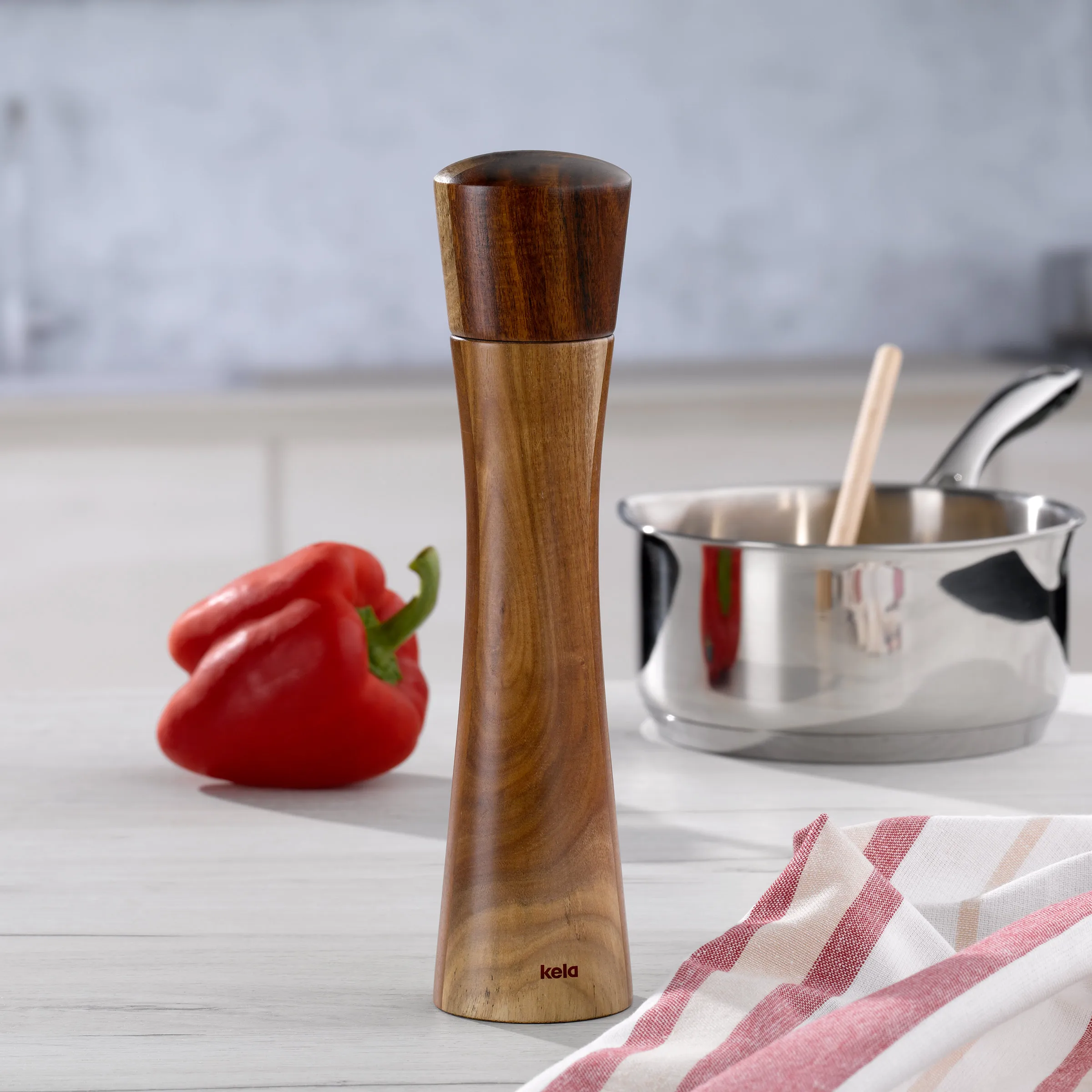 Holz-Pfeffermühle Kela - Elegante Gewürzmühle für die Küche Holz-Pfeffermühle von Kela auf Küchenarbeitsfläche mit roter Paprika und Kochtopf im Hintergrund