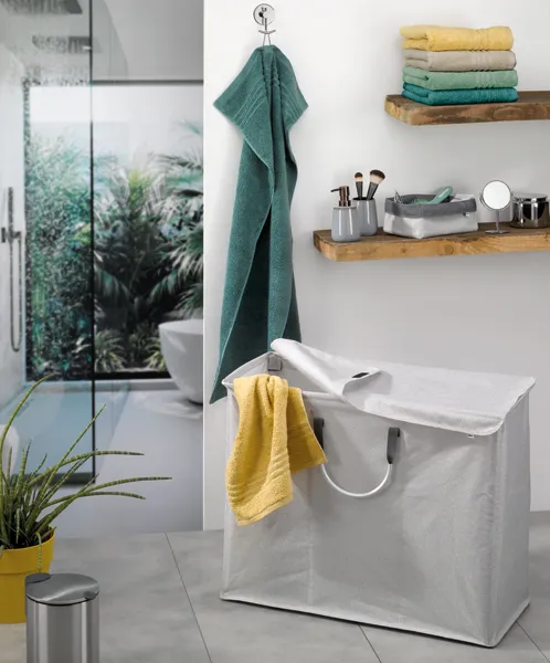 Modernes Badezimmer mit grünem Handtuch, Wäschekorb, Holzregalen, Kosmetikprodukten und begehbarer Dusche mit Pflanzen