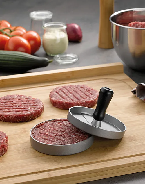 Burger-Presse zum Formen von Fleischpatties - Profi-Küchenzubehör Burger-Presse mit rohem Fleischpatty auf Holzbrett, umgeben von Tomaten, Zwiebeln und Zutaten in Schüsseln.