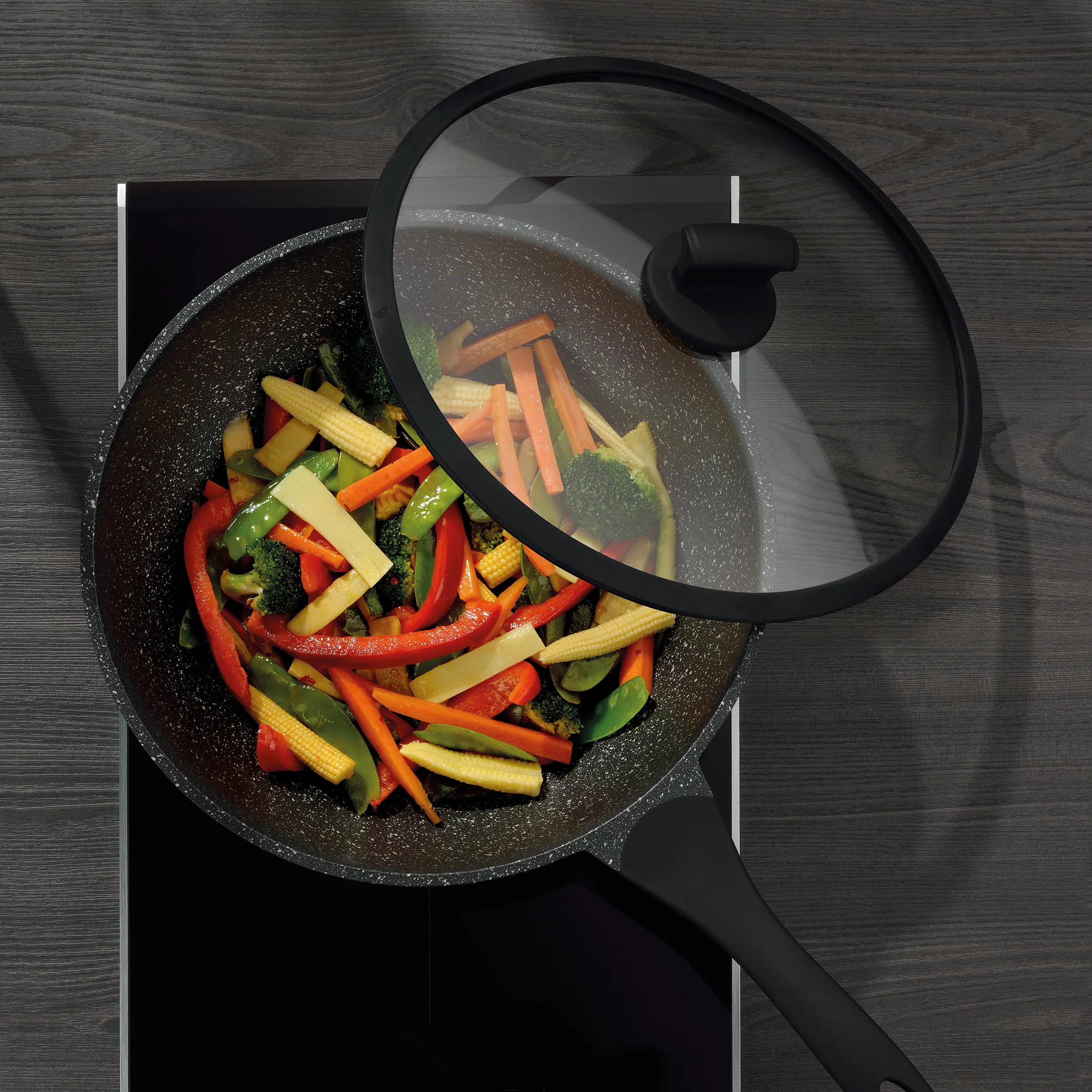 Wok-Pfanne mit Gemüse und Glasdeckel Wok-Pfanne mit Deckel auf schwarzem Kochfeld, gefüllt mit sautiertem Gemüse wie Brokkoli, Paprika, Mais und grünen Bohnen