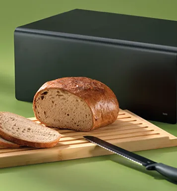 Vollkornbrot Brotkasten - frisches Brot lagern Frisches Vollkornbrot mit Schnittmuster auf Holzbrett neben schwarzem Brotkasten und grünem Hintergrund