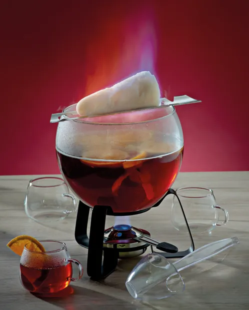 Feuerzangenbowle Punsch - traditionelle Bowlenzubereitung mit Flamme Glühender Punsch in einer Feuerzangenbowle mit brennendem Zuckerhut, umgeben von Gläsern und rotem Hintergrund