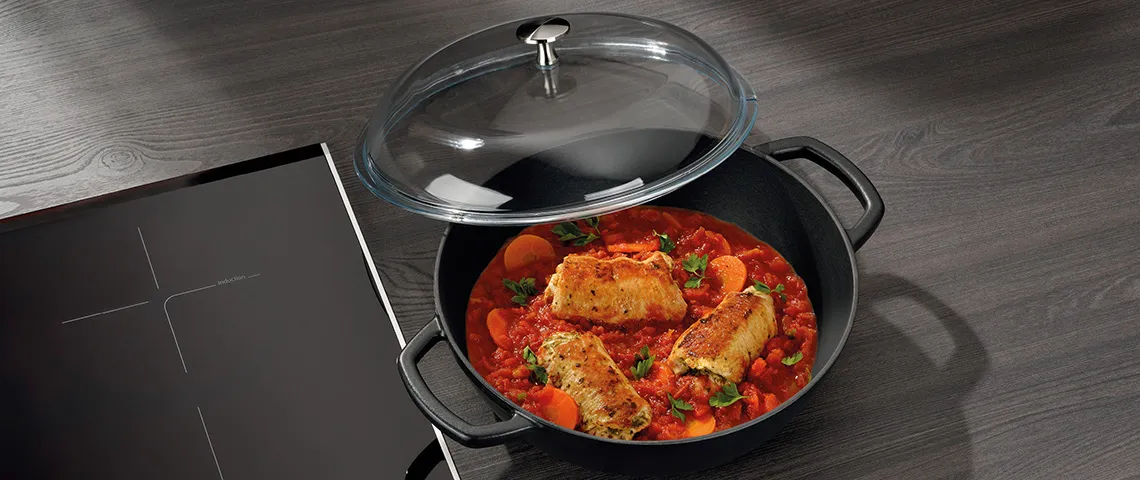 Bräter Induktion mit Glassdeckel - Hähnchen Schmortopf Schwarzer Bräter mit Glassdeckel auf Induktionskochfeld, darin Hähnchenschenkel in roter Tomatensauce mit Gemüse