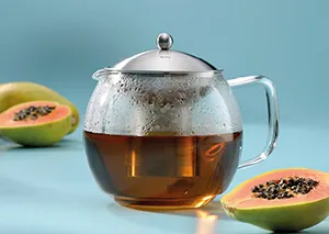 Glas-Teekanne mit Sieb und frischem Früchtetee - Teezubereitung Glas-Teekanne mit Teesieb gefüllt mit goldenem Tee, daneben aufgeschnittene Papayas auf blauem Hintergrund
