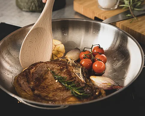 Steak mit Kräutern und Gemüse in einer Edelstahlpfanne mit Holzspatel, umgeben von Tomaten und Rosmarin.