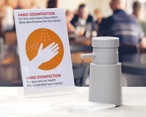 Handdesinfektionsspender mit zweisprachigem Hinweisschild für Handhygiene auf weißem Tisch