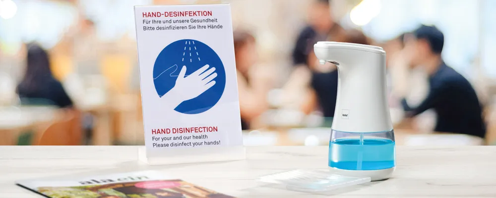 Automatischer Handdesinfektionsspender mit blauer Flüssigkeit neben Hinweisschild zur Handhygiene am Empfang