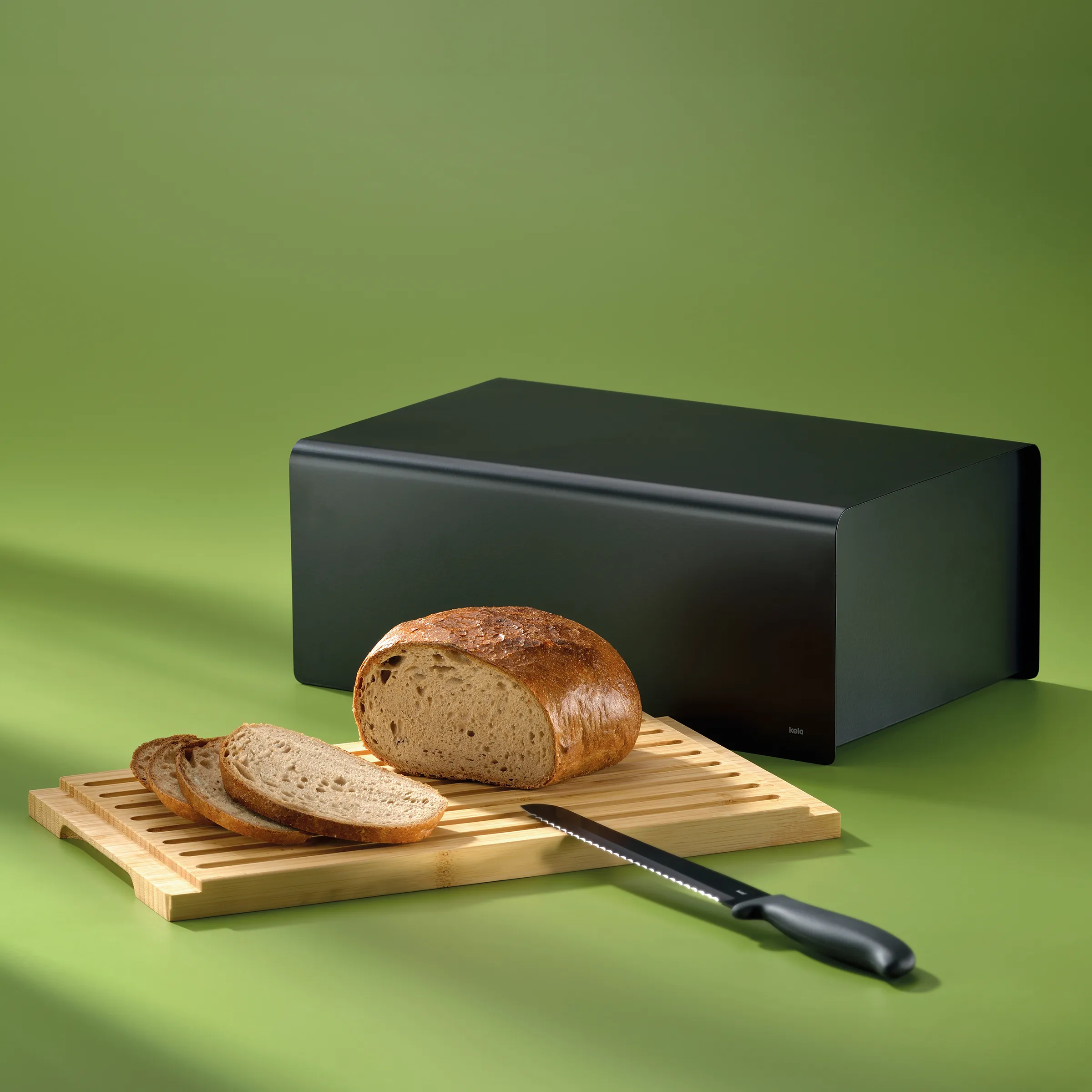 Brotkasten schwarz - moderne Brotaufbewahrung Schwarzer Brotkasten aus Metall neben Holzbrett mit geschnittenem Vollkornbrot und Brotmesser auf grünem Hintergrund