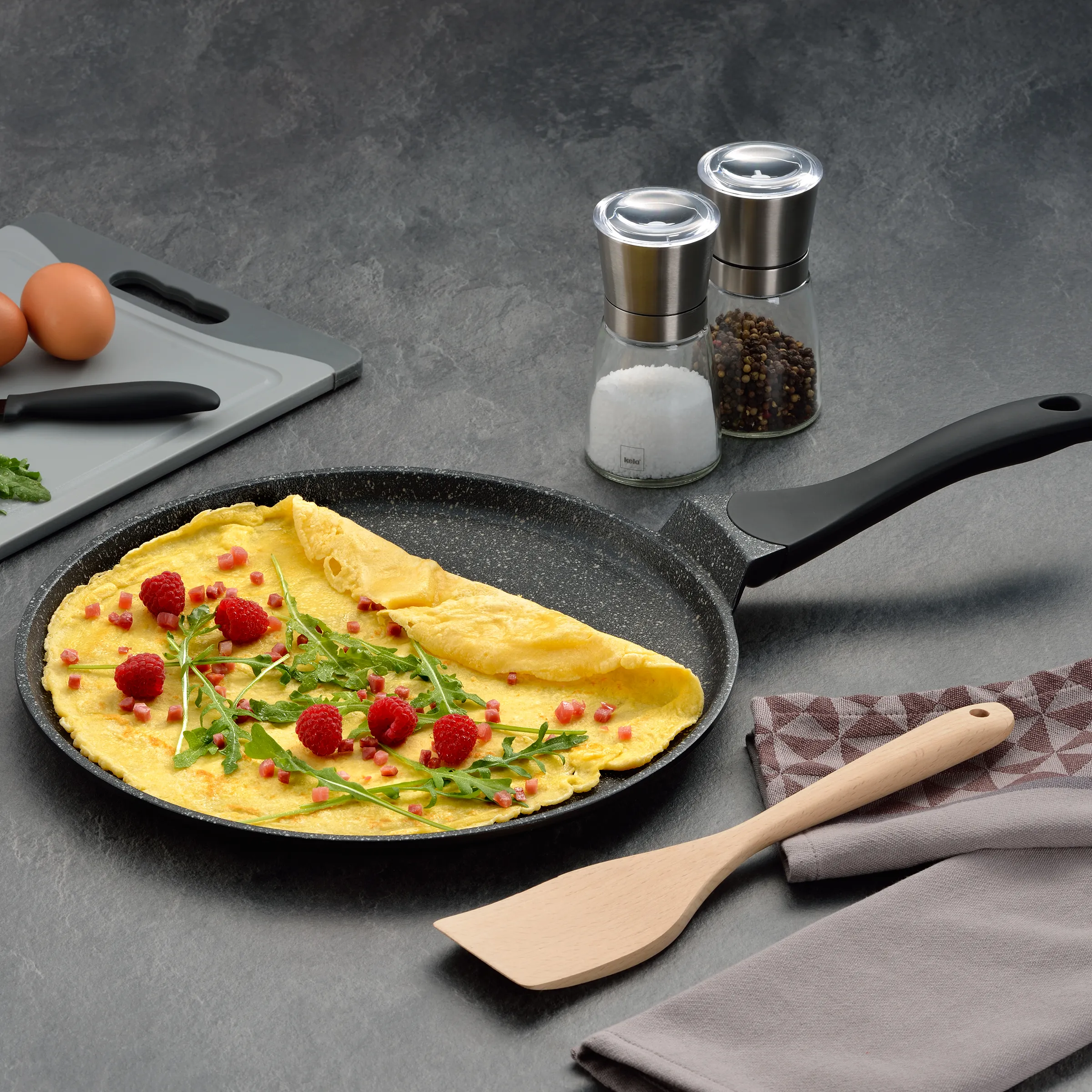 Omelette Rezept: Fluffy Omelett mit Himbeeren zubereiten Fluffy Omelette in schwarzer Pfanne mit Himbeeren, Granatapfelkernen und Rucola auf grauem Untergrund