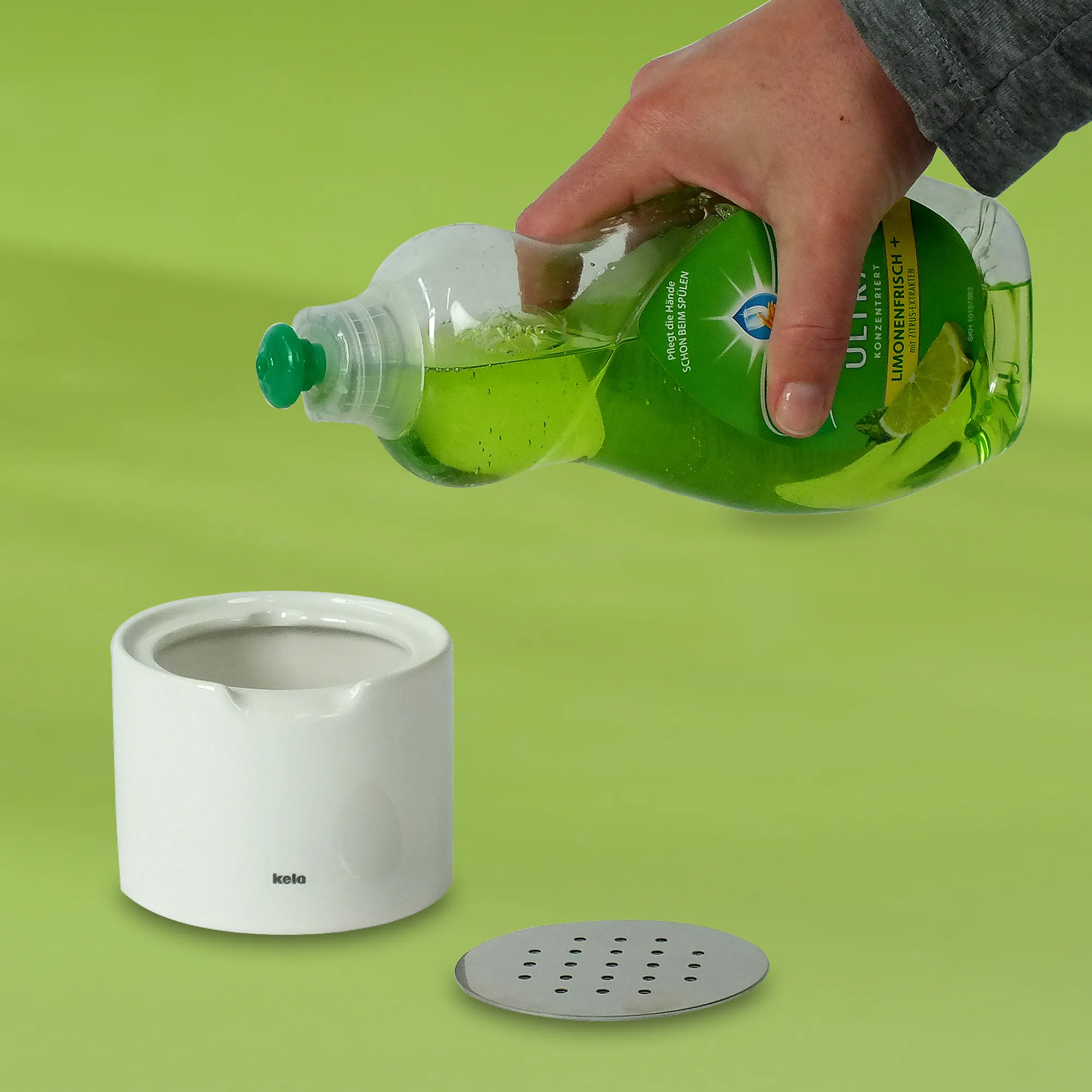 Nachfüllbarer Seifenspender mit Spülmittel befüllen - Kela Bad-Accessoires Hand gießt grüne Spülmittel-Flasche in weißen Kela Seifenspender mit Lochsieb auf grünem Hintergrund