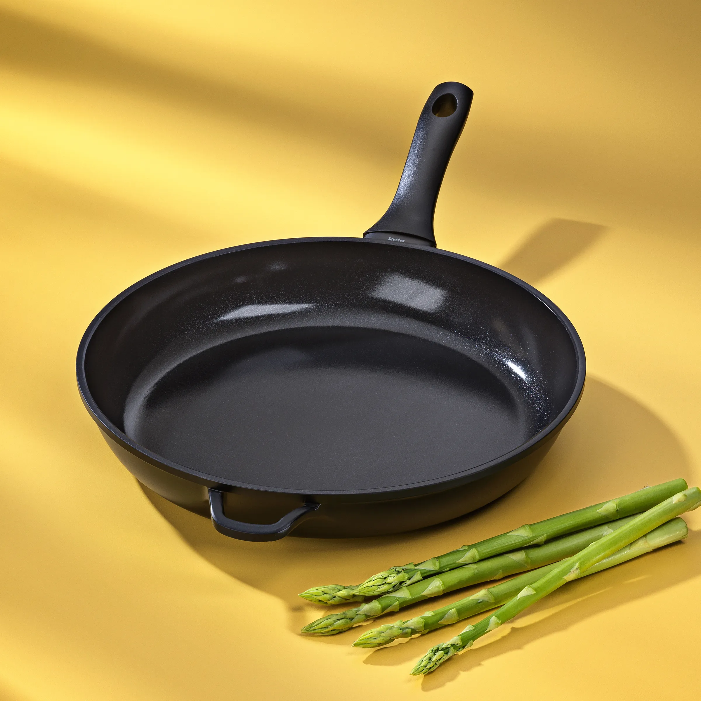 Bratpfanne Antihaftbeschichtung Induktion Küche Schwarze Bratpfanne mit Griff auf gelbem Hintergrund, daneben frischer grüner Spargel