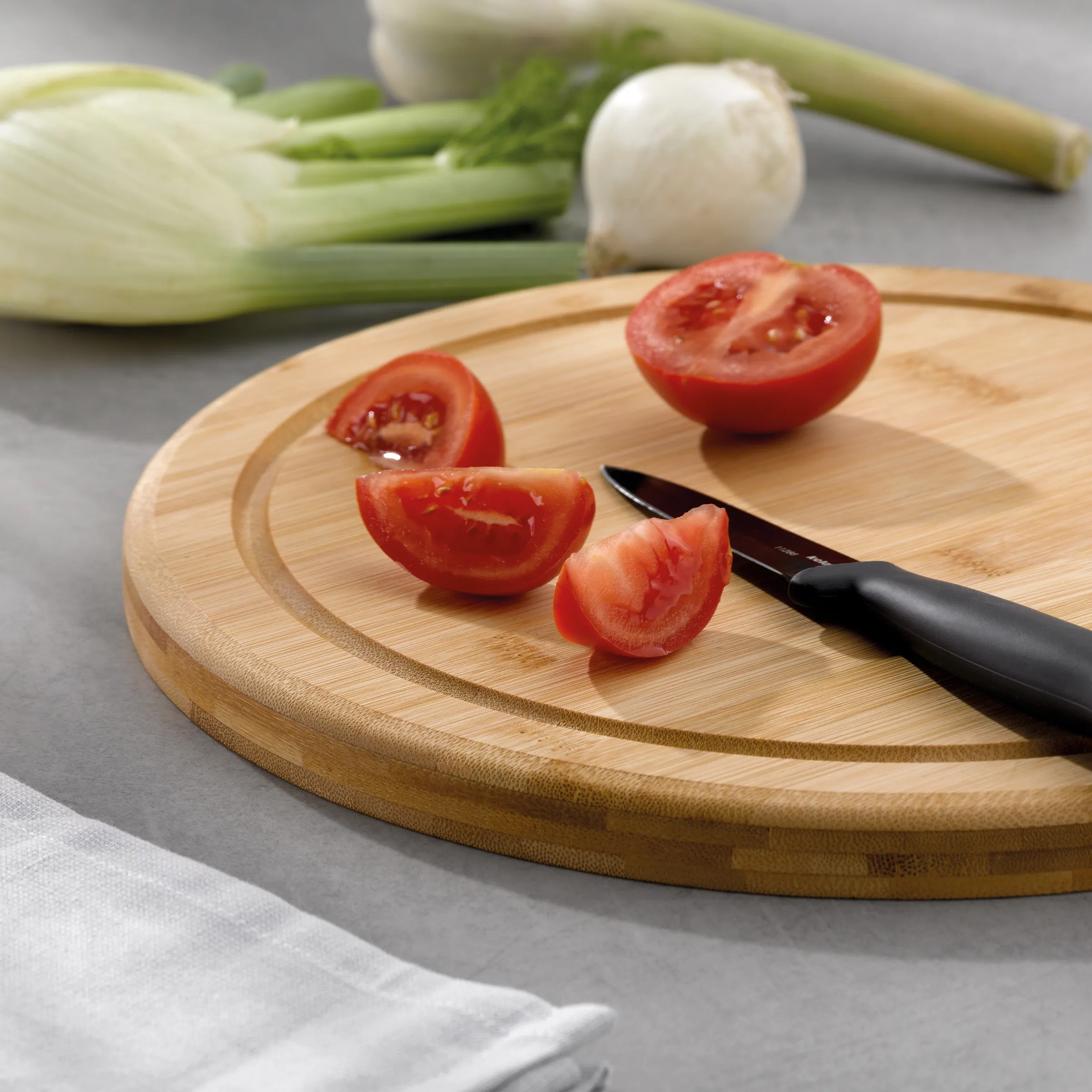 Tomaten schneiden auf Schneidebrett - frische Zutaten für Küche Frische rote Tomaten werden auf einem runden Bambusschneidebrett mit schwarzem Messer geschnitten. Im Hintergrund liegen Lauch und Knoblauch.