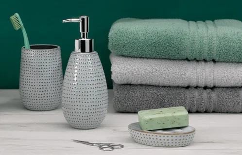 Badezimmer-Set mit grauem Seifenspender, Zahnbürstenbecher, gestapelten Handtüchern, Seife und Nagelschere auf weißem Untergrund vor grünem Hintergrund.