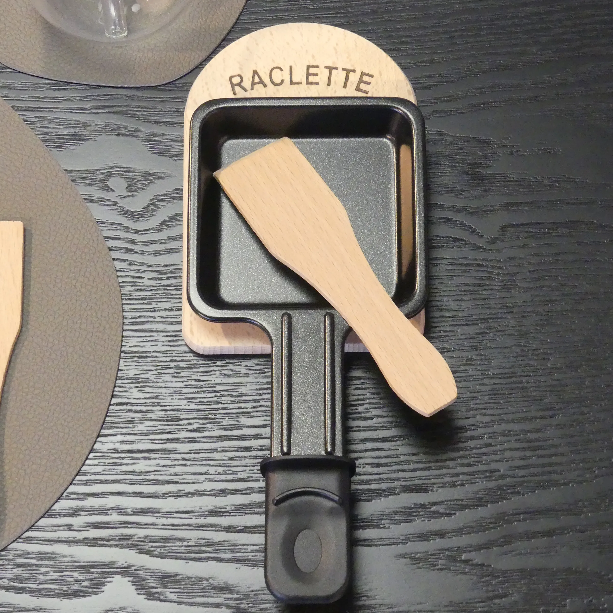 Raclette-Spachtel Holz - Zubehör für Raclette-Pfanne Raclette-Spachtel aus Holz mit schwarzem Griff auf Raclette-Pfanne mit grauer Oberfläche auf dunklem Holztisch