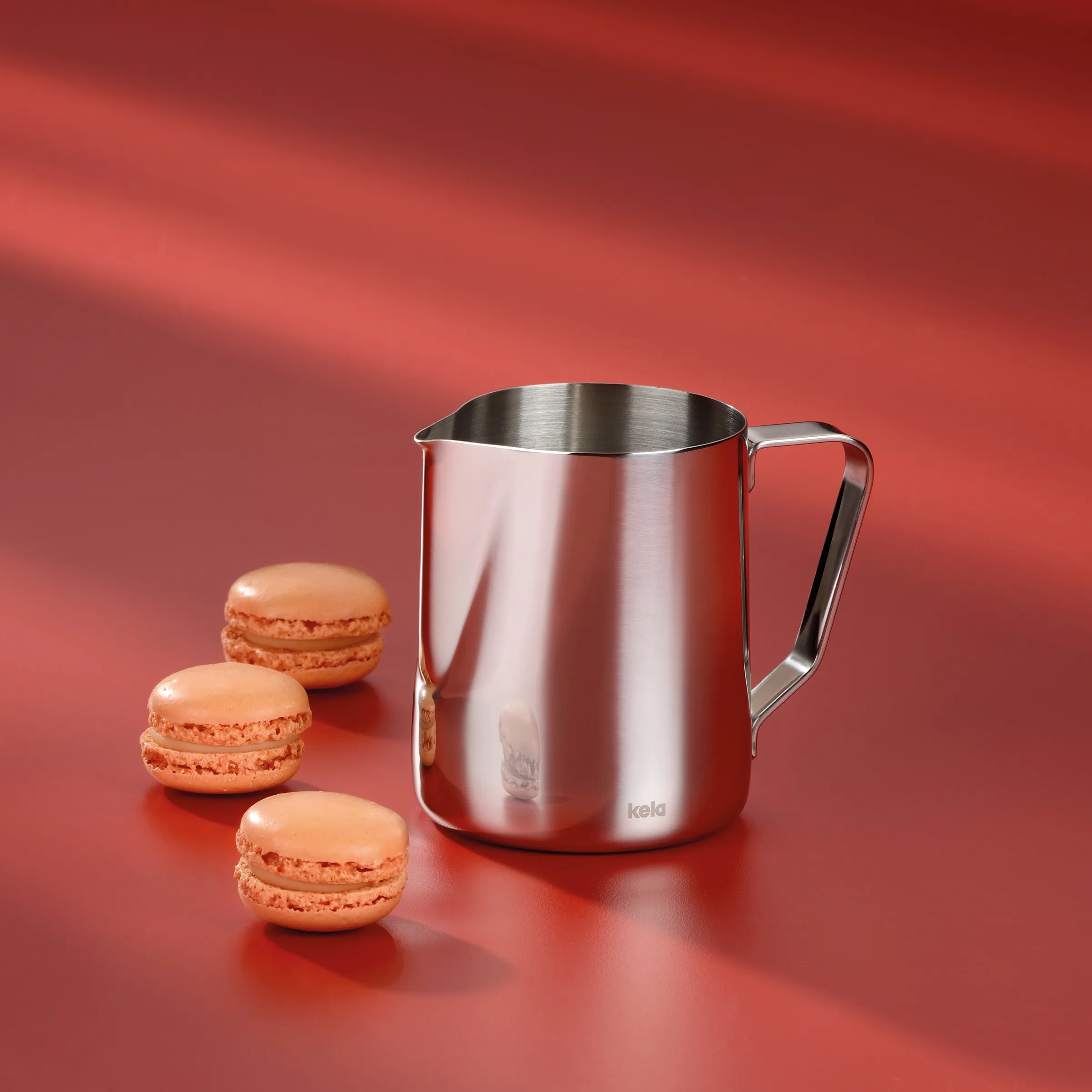 Kela Milchkännchen Edelstahl mit Macarons - Barista-Zubehör Edelstahl-Milchkännchen der Marke kela mit drei rosa Macarons vor rotem Hintergrund