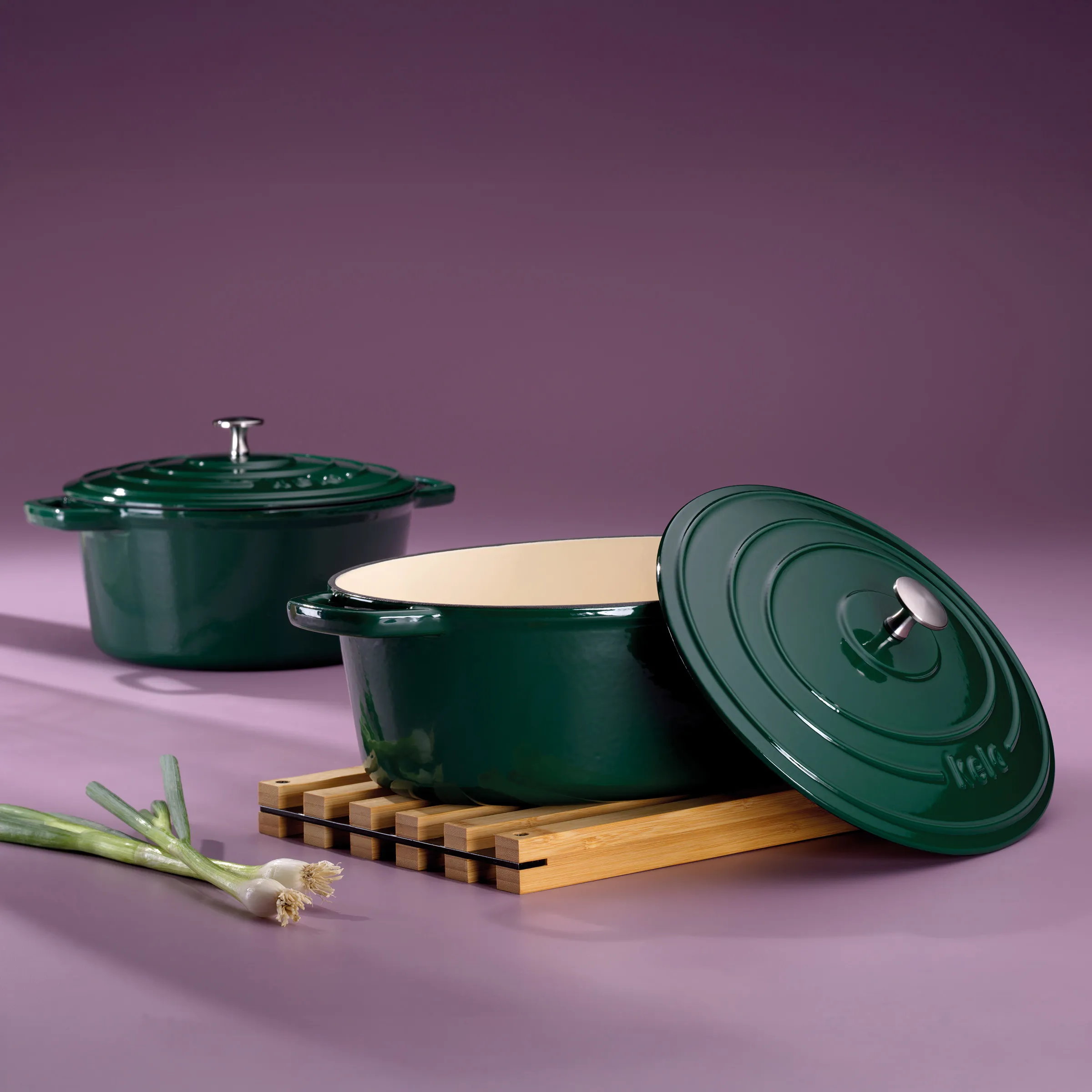 Zwei dunkelgrüne gusseiserne Bräter mit Deckel auf Holzuntersetzer, daneben Frühlingszwiebeln