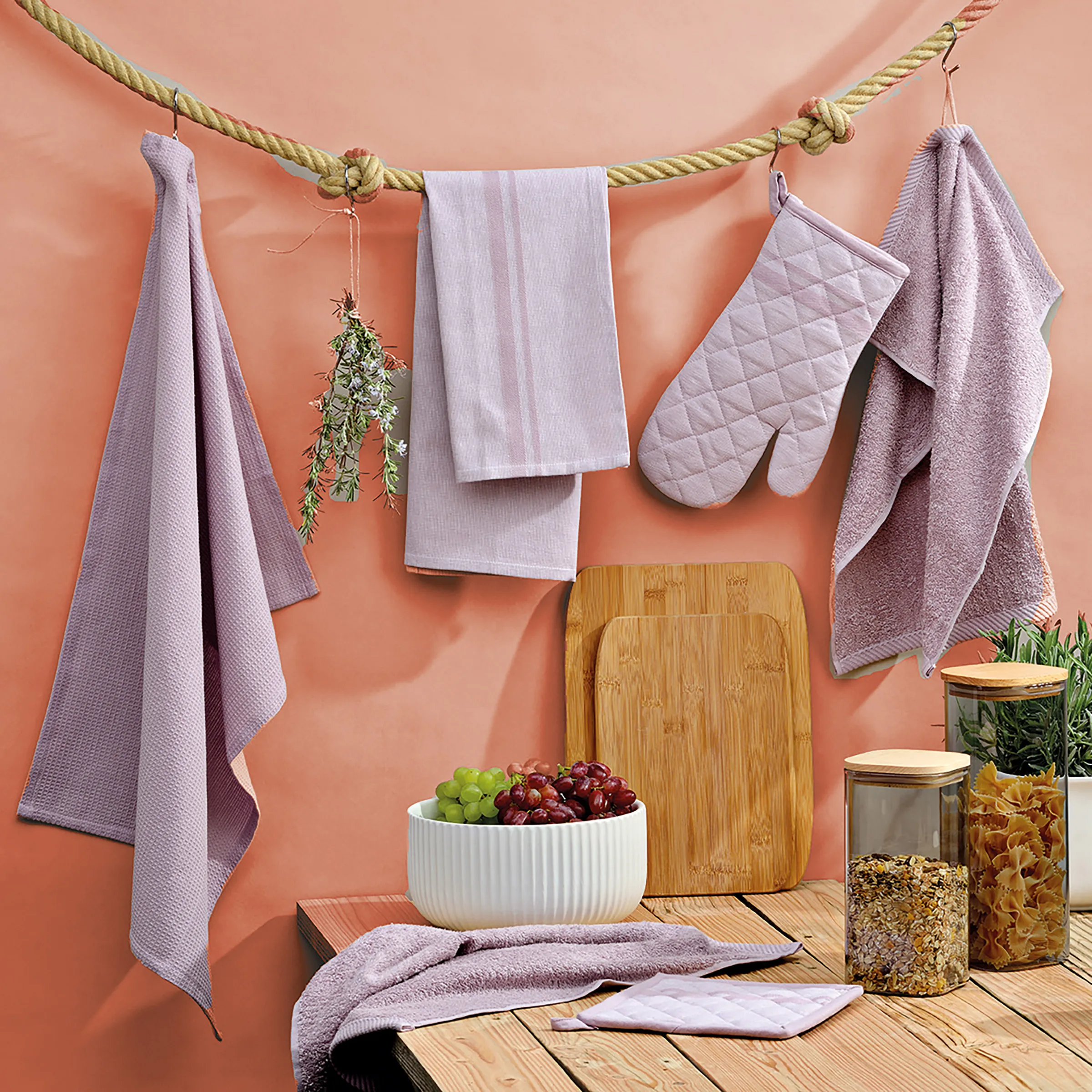 Küchenhandtücher und Küchenzubehör Set - Lila Textilien Küchenaufbau mit lila Handtüchern an Seil, Oven-Handschuh, Holzbretter, Schüssel mit Trauben und Gewürzgläsern vor korallefarbenem Hintergrund