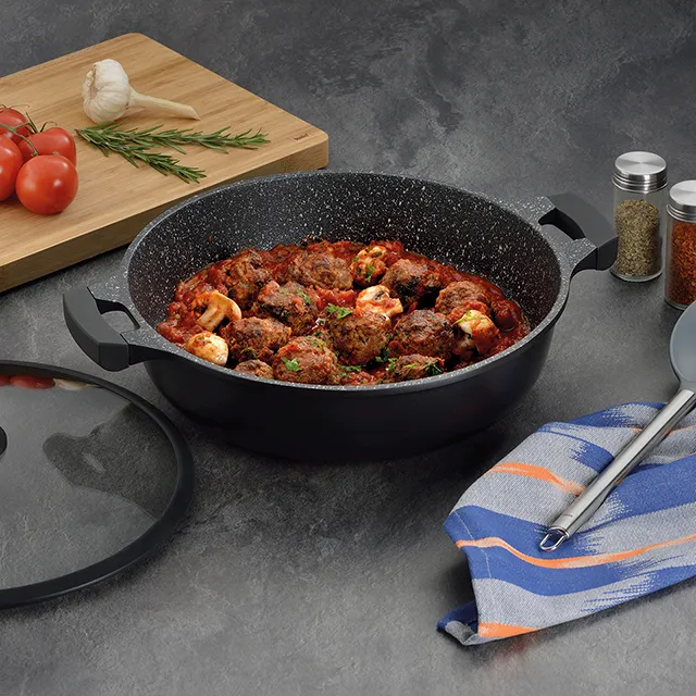 Wok mit Fleischbällchen - Rezept und Zubereitung Schwarzer Wok mit Fleischbällchen in Tomatensauce auf grauer Oberfläche, daneben Schneidebrett mit Tomaten, Knoblauch und Rosmarin