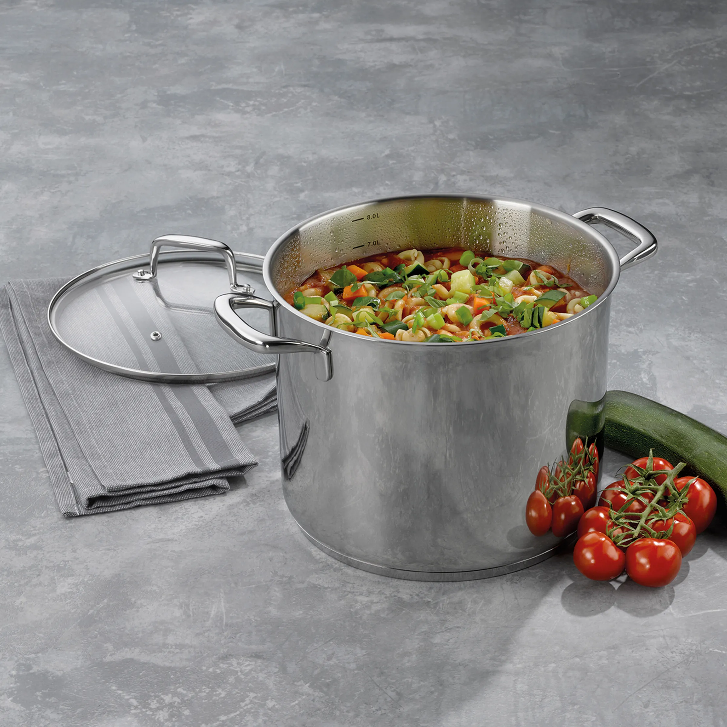 Edelstahl-Kochtopf mit Glasdeckel und Gemüsesuppe - Premium Kochgeschirr Edelstahl-Kochtopf mit Glasdeckel, gefüllt mit Gemüsesuppe, neben Kirschtomaten und Zucchini auf grauer Oberfläche