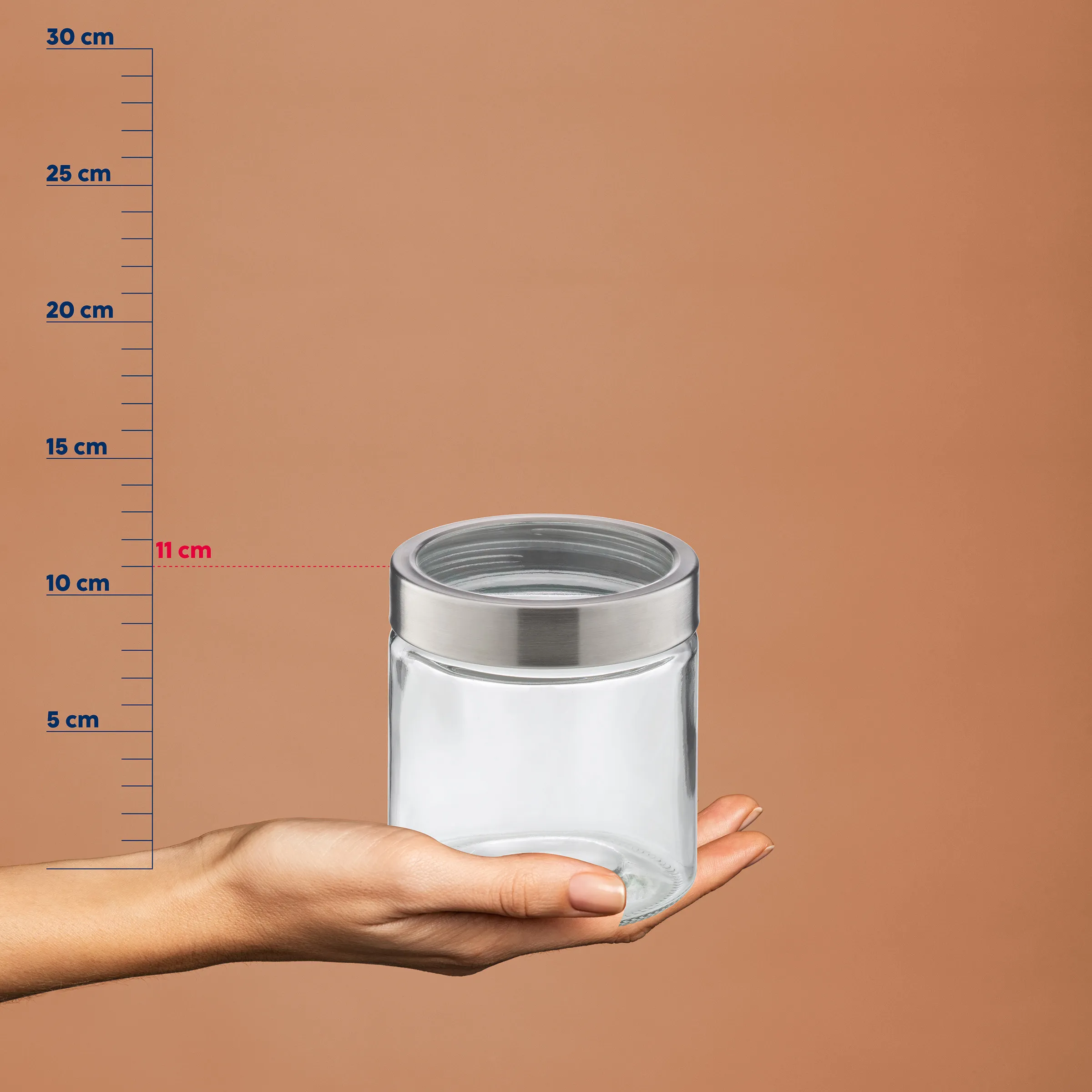 Hand hält transparentes Vorratsglas mit Metalldeckel, Höhe 11 cm, vor Maßstab auf beigem Hintergrund
