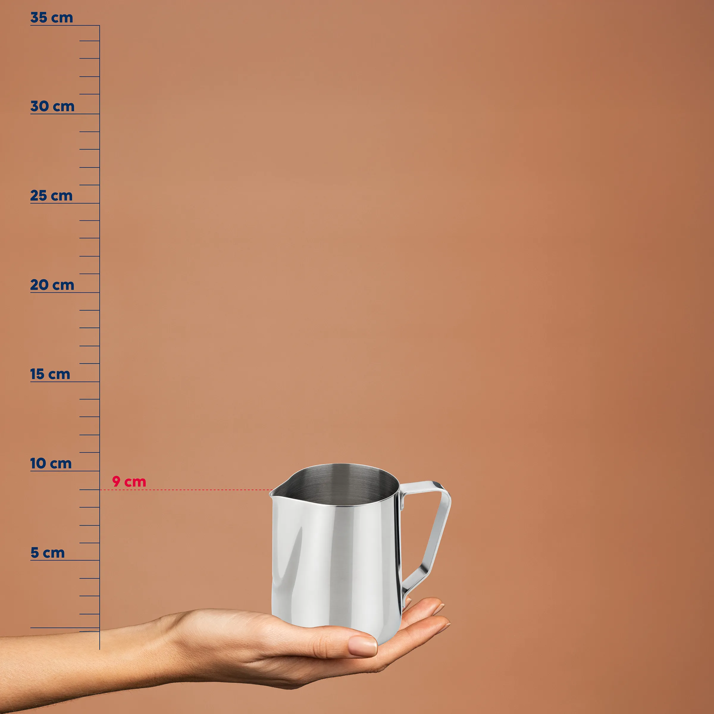 Milchkännchen Edelstahl - Professionelle Barista-Kanne Edelstahl-Milchkännchen mit Henkel in einer Hand vor einer Messskala, zeigt 9 cm Höhe