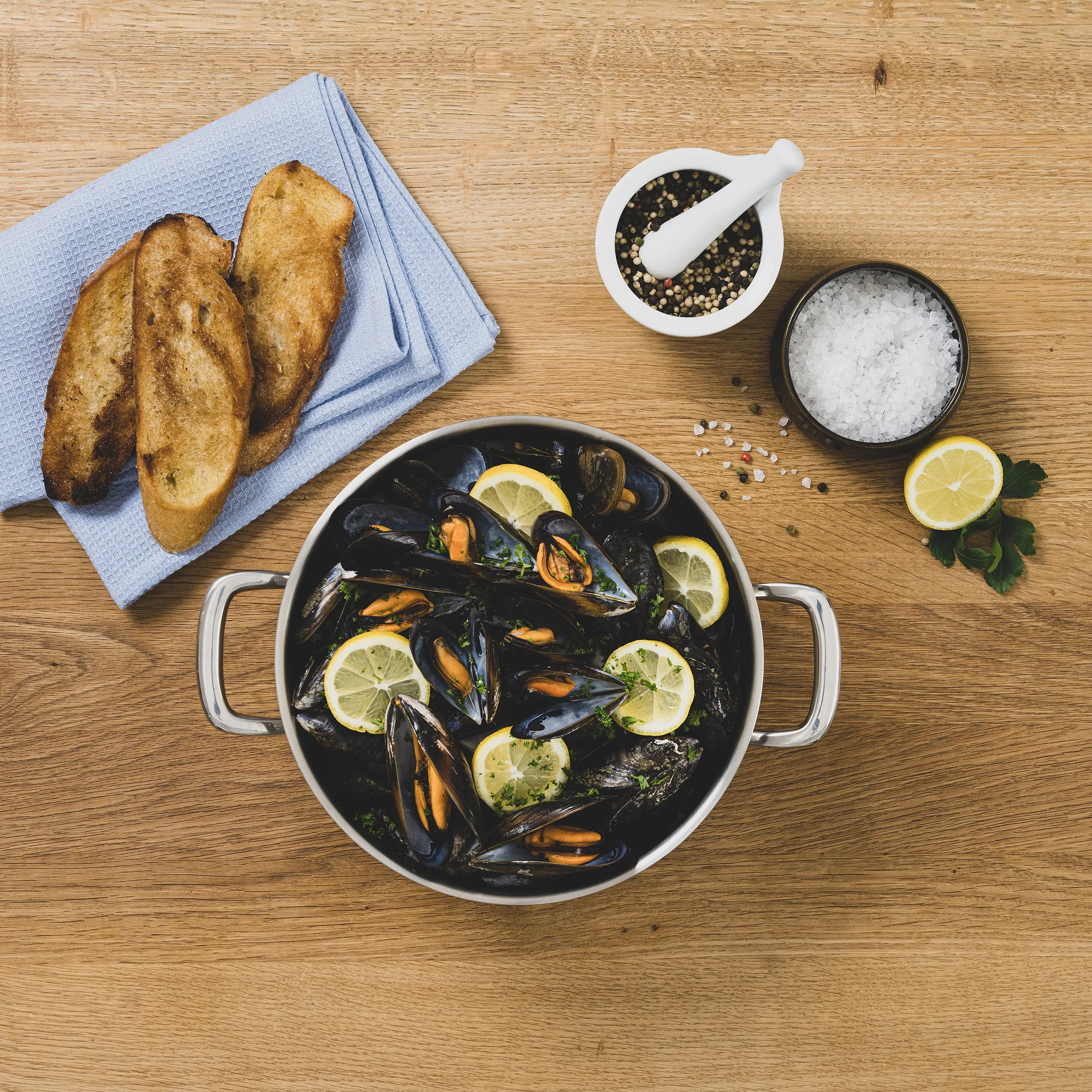 Muscheln Rezept - Klassische Meeresfrüchte Zubereitung Muscheln mit Zitrone in weißem Topf, Brot auf blauem Tuch, Salz, Pfeffer und Zitrone auf Holztisch