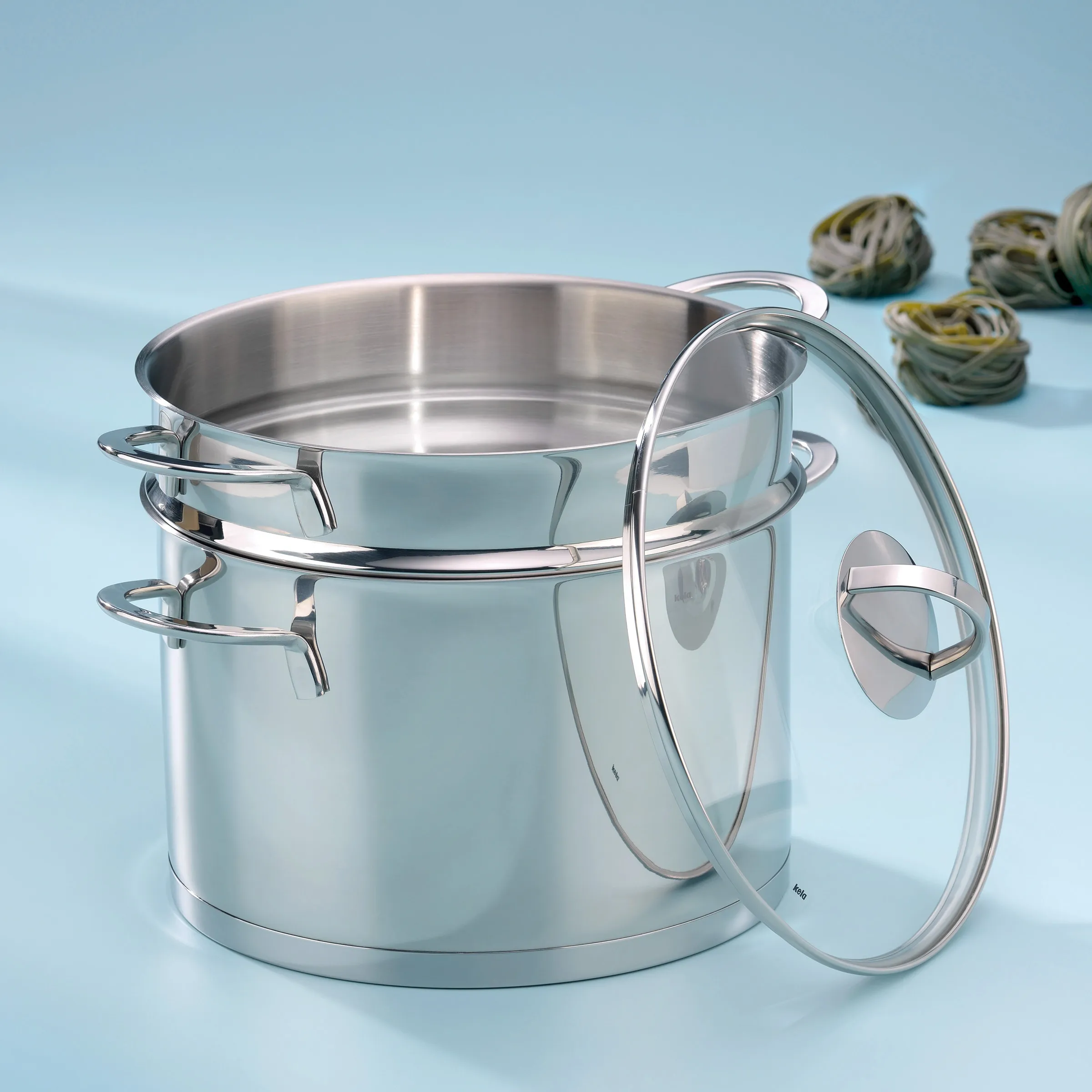 Edelstahl Kochtopf mit Glasdeckel - hochwertiges Kochgeschirr Edelstahl-Kochtopf mit Glasdeckel und Metallgriffen auf hellblauem Hintergrund, daneben getrocknete Artischocken