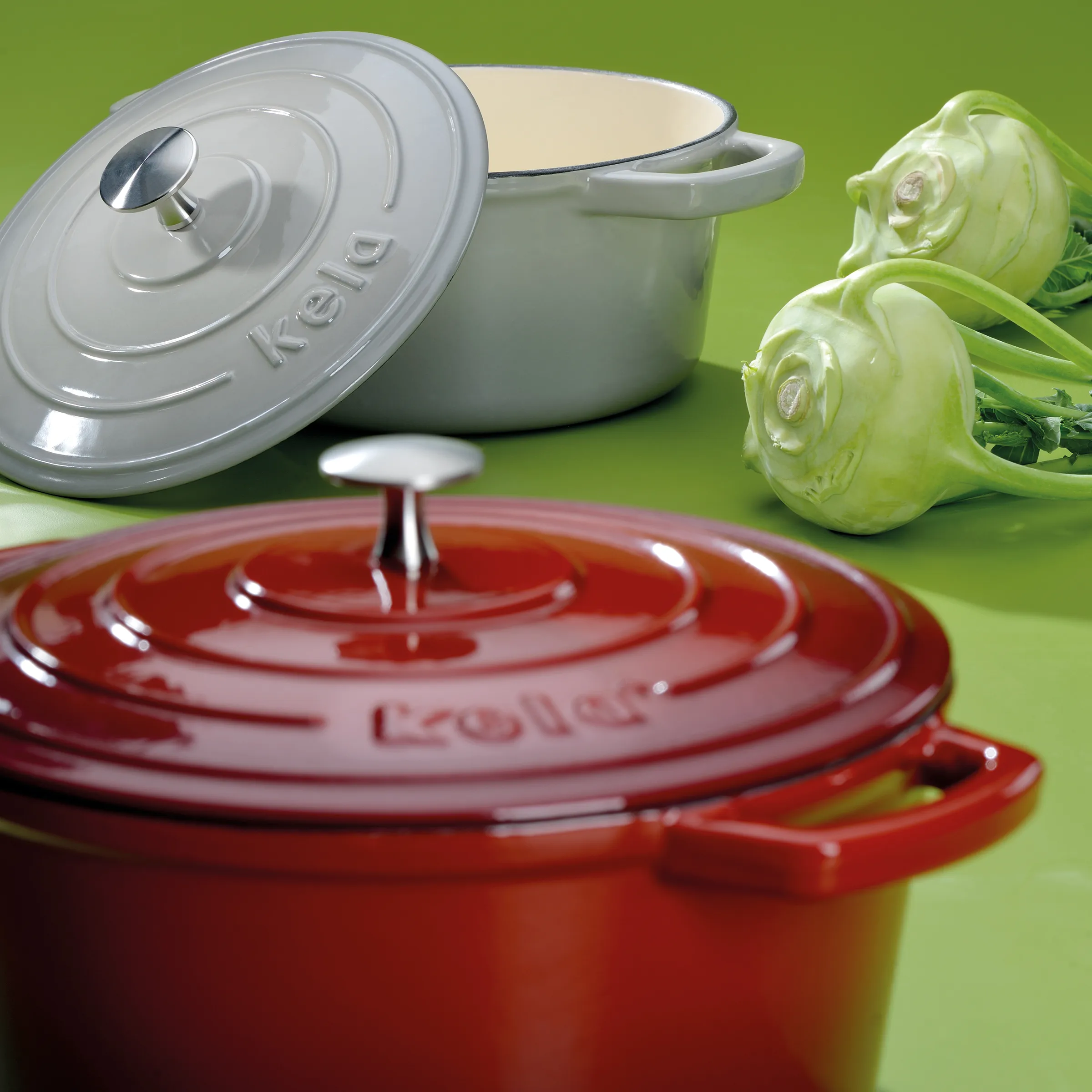 Rote und graue Kela Gusseisen-Kochtöpfe mit Deckeln und frischem Kohlrabi auf grünem Hintergrund